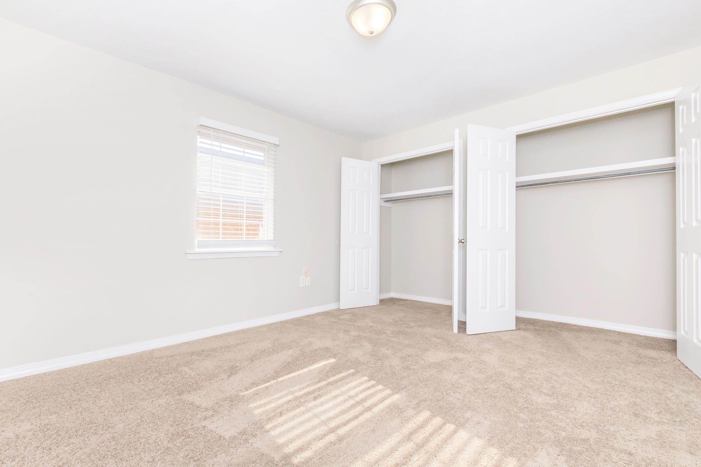 Empty room with beige walls and carpet, featuring two open closet doors. Natural light streams in through a window with white blinds, highlighting the clean and simple design of the space.
