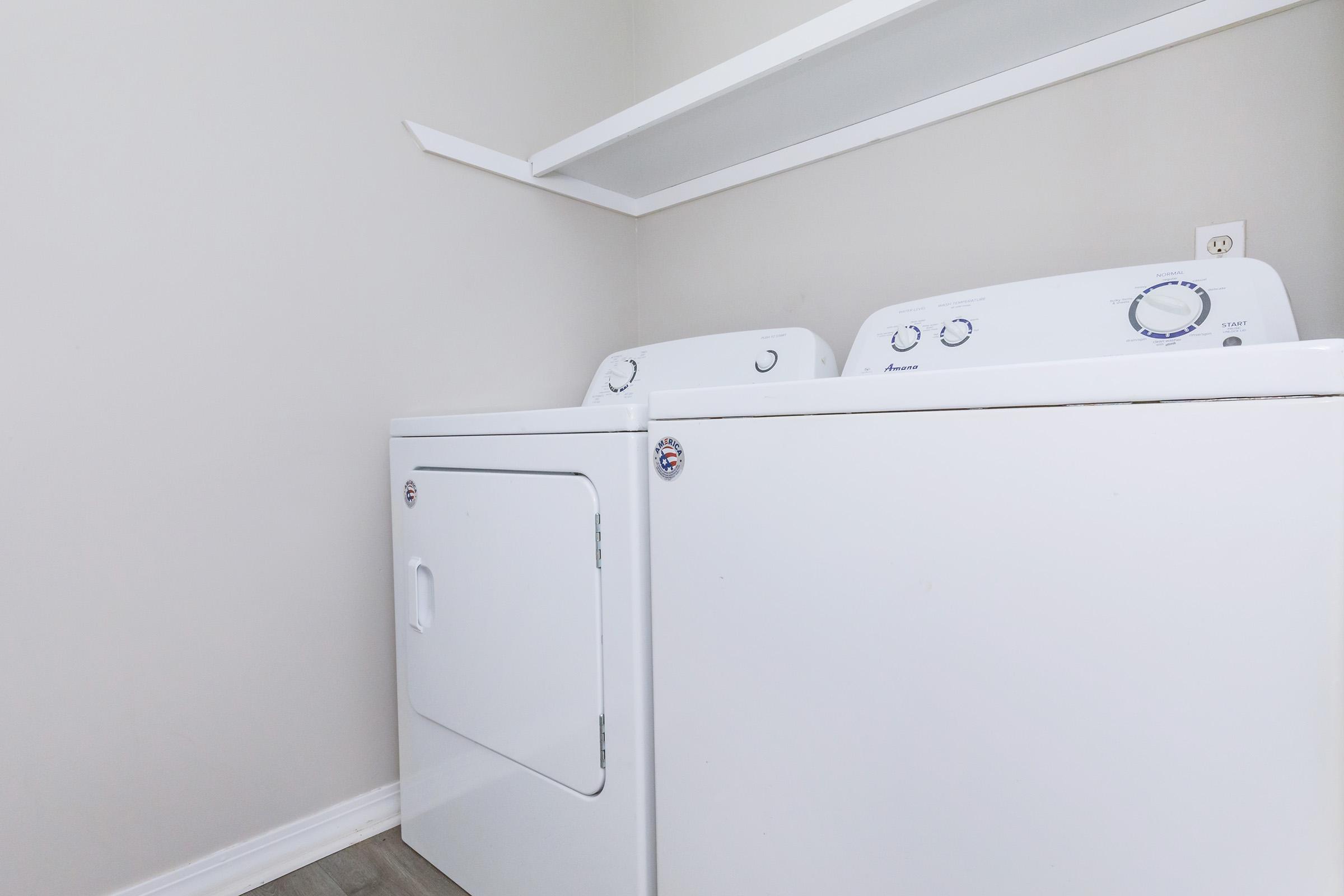 A clean laundry room featuring a white washing machine and dryer side by side, with a simple white shelf above. The walls are painted in a light gray color, and the floor is a light wood finish, creating a bright and organized space for laundry tasks.