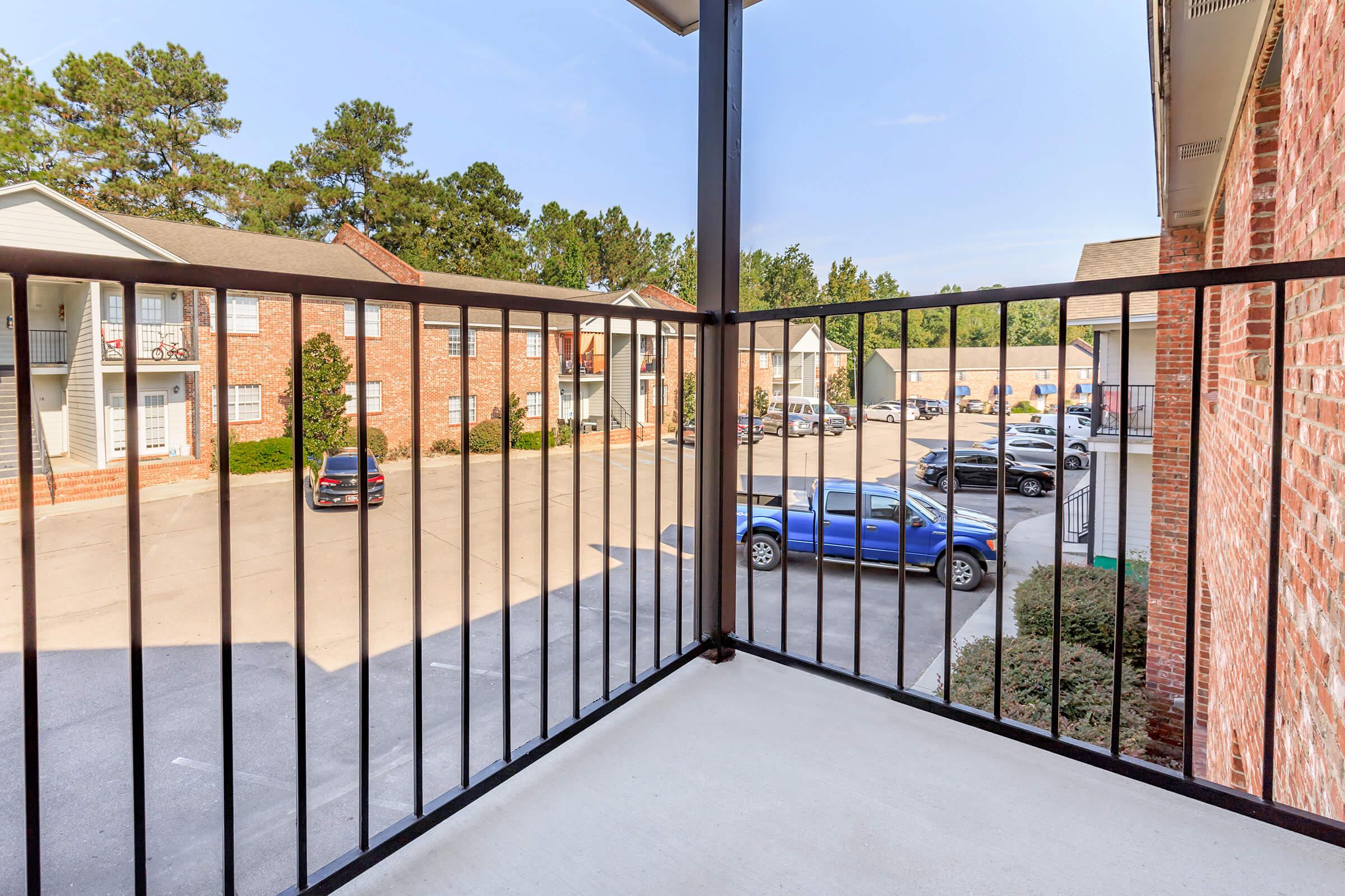View from a balcony overlooking a parking lot with several parked cars and nearby residential buildings surrounded by trees. The scene features a clear sky and a well-maintained outdoor area.
