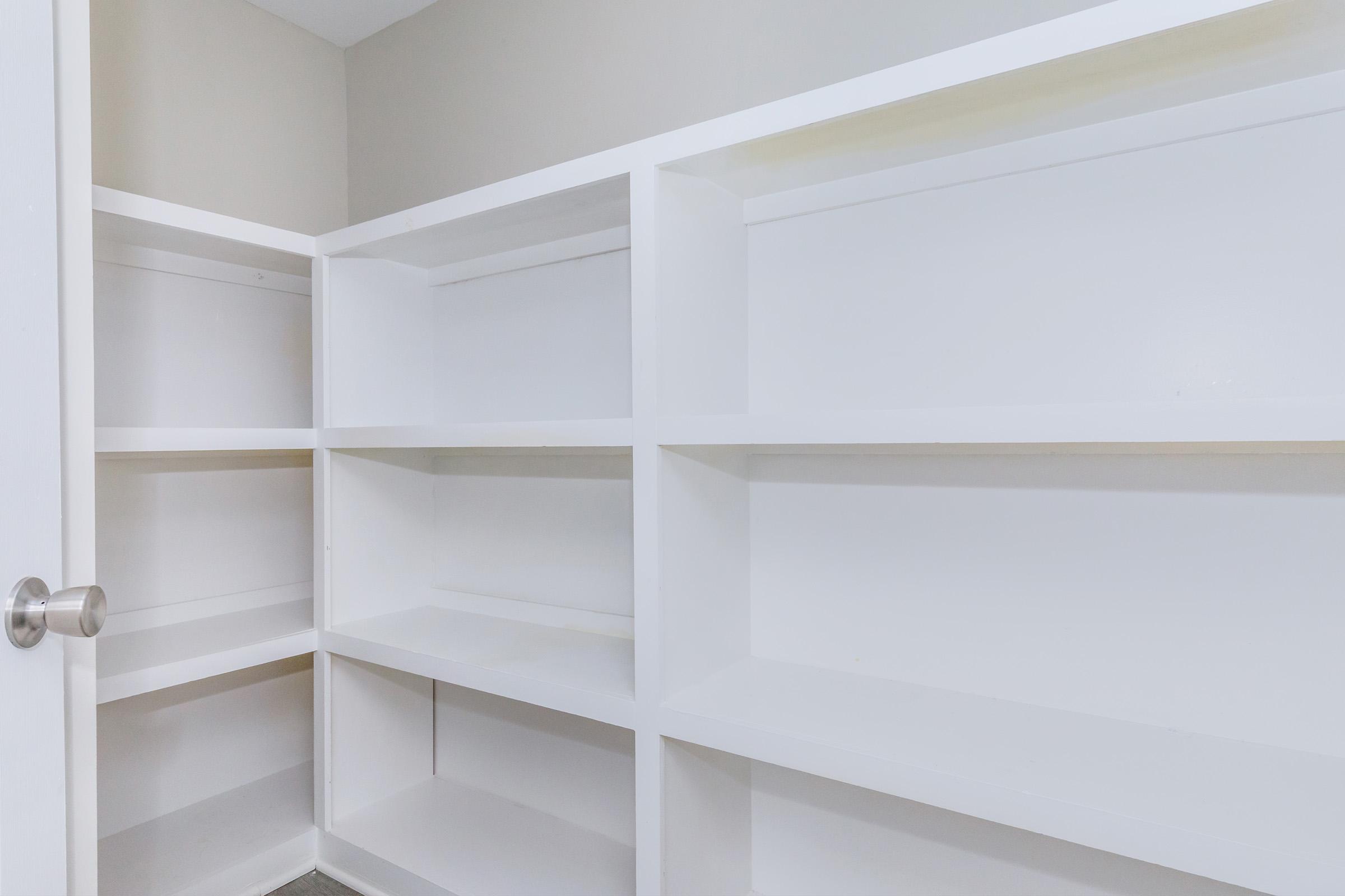 Empty white shelving unit against a wall, featuring multiple flat shelves with no items displayed. The background is a light beige color, and a door handle is visible on the left side of the image. The overall scene conveys a minimalist and tidy storage space.
