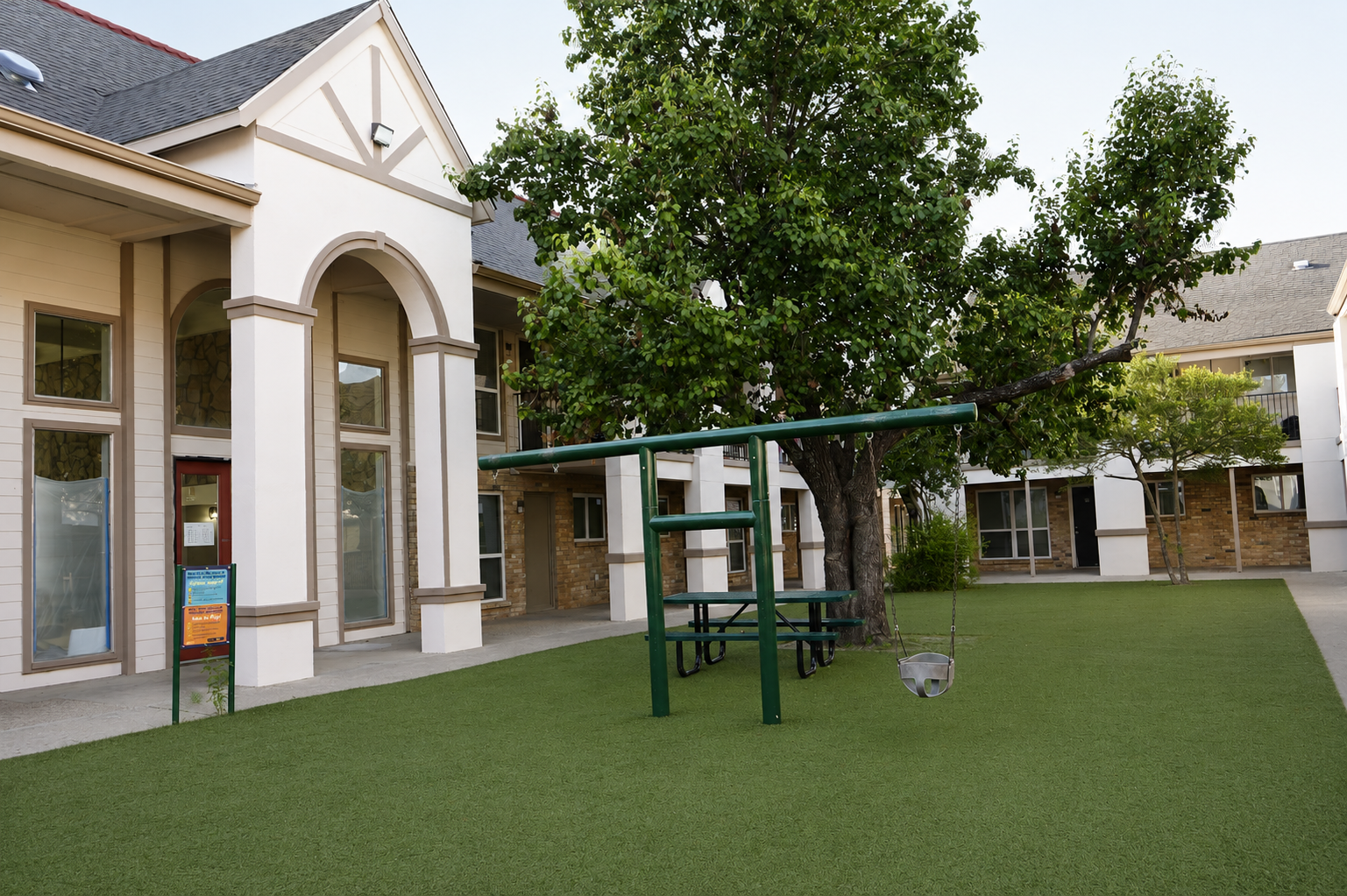 A grassy courtyard featuring a tree, a swing set, and a picnic area with tables. The building in the background has large windows and a welcoming entrance. The space is designed for relaxation and play, with synthetic grass providing a neat, green environment.