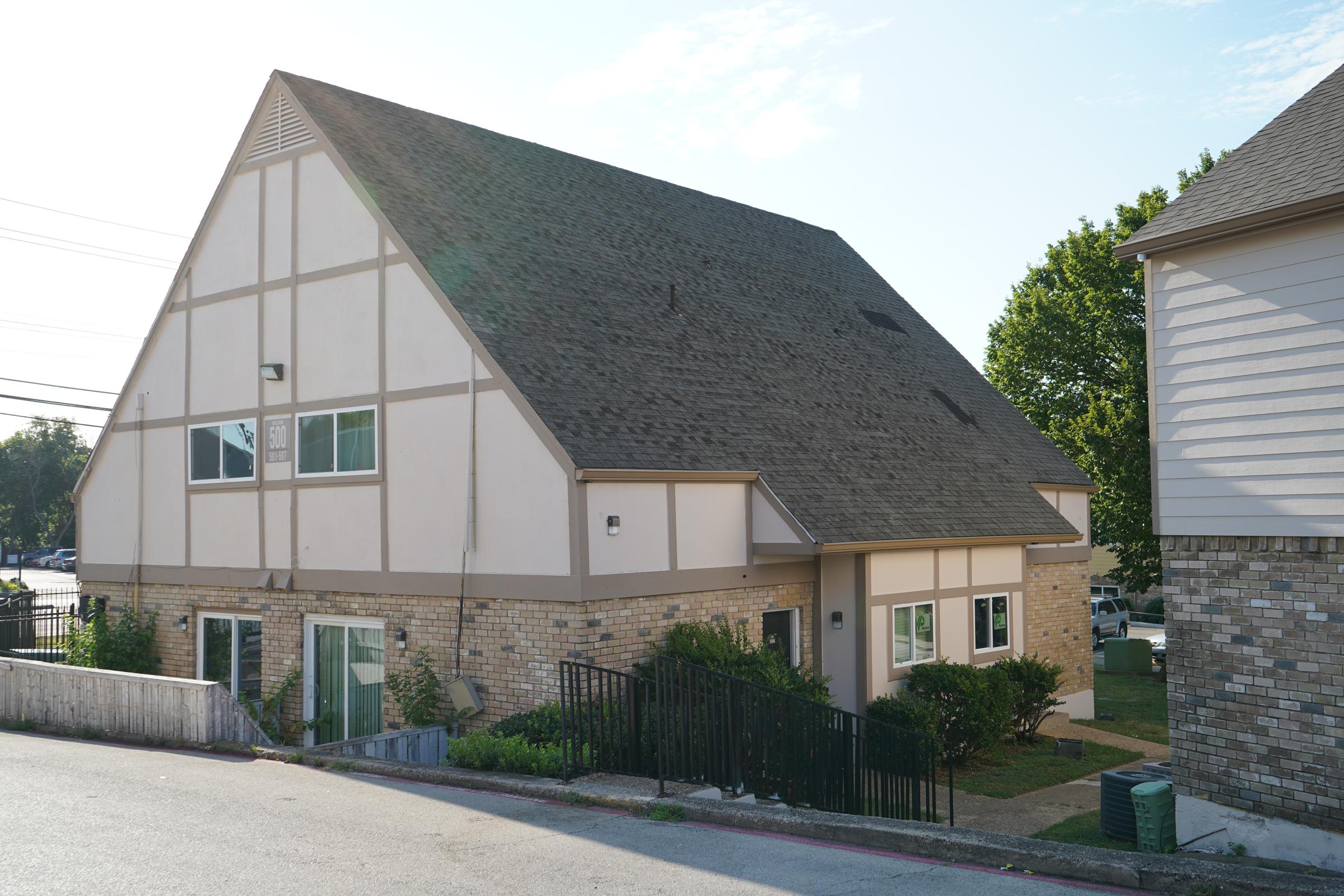A two-story building with a sloped roof, featuring a mix of stone and pale exterior siding. It has multiple windows and a small garden area, surrounded by a grassy lawn. The setting includes a nearby sidewalk and a tree, with power lines visible in the background under a clear blue sky.