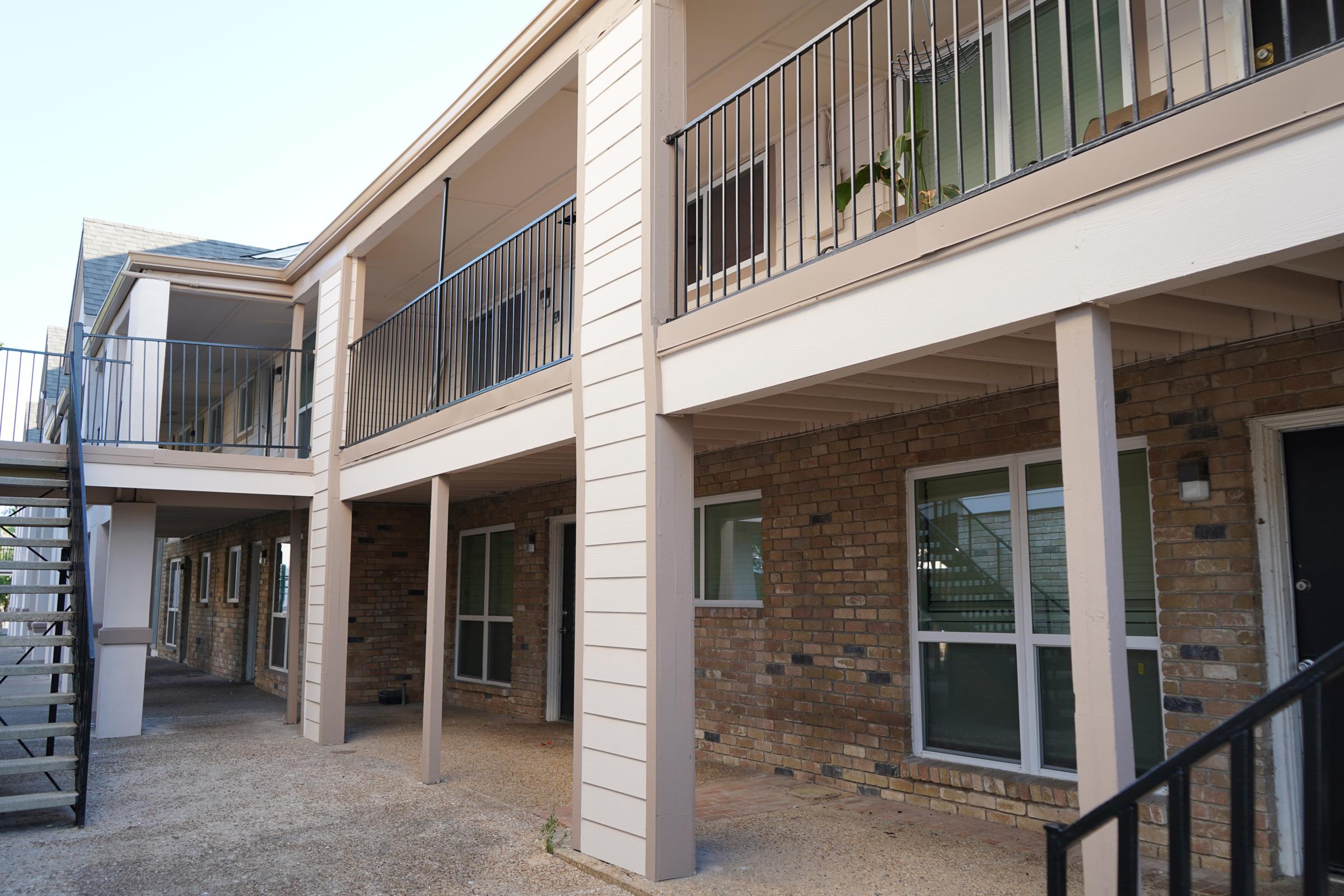 Exterior view of a multi-story apartment building featuring a staircase and balconies. The facade is a combination of brick and light-colored siding, with multiple windows visible on each floor. The area is well-lit, and there are some potted plants on the balconies.