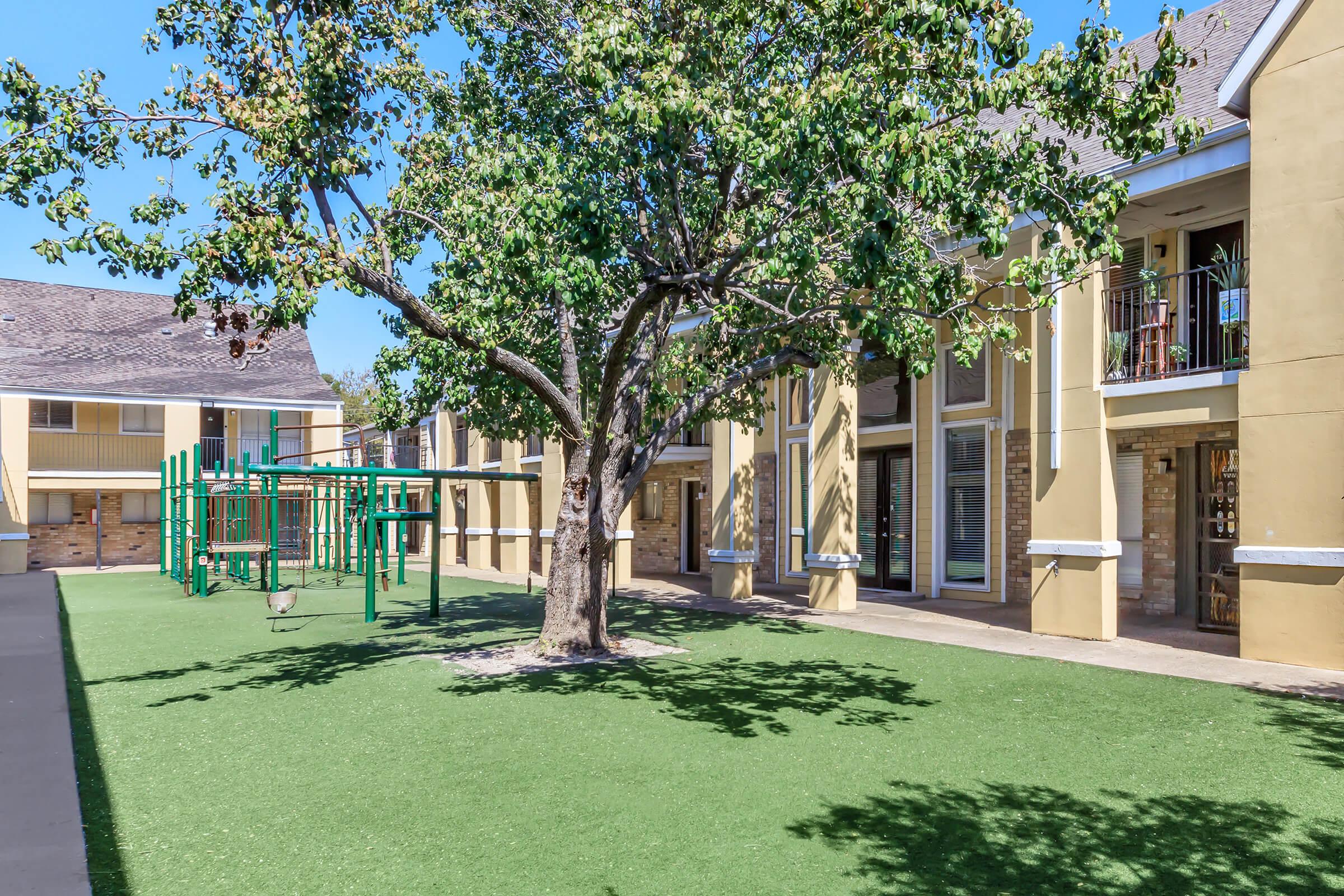A green courtyard featuring a large tree and a playground with swings and climbing structures. Surrounding the area are residential buildings with balconies, all under a clear blue sky. The grass is well-maintained, creating a welcoming outdoor space for recreation and relaxation.