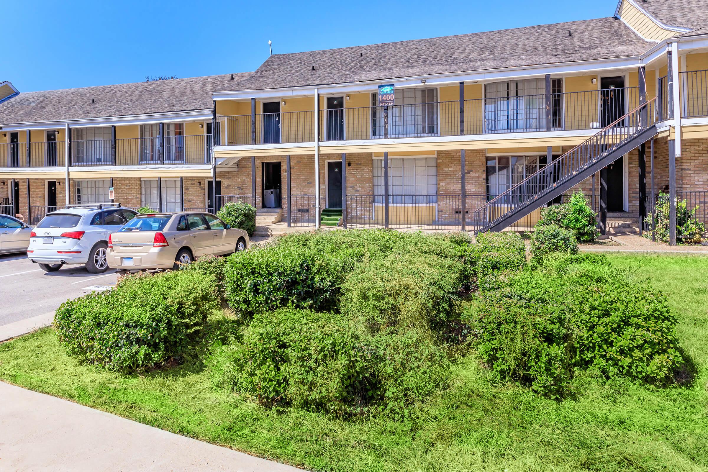 A view of a multi-story apartment building with yellow siding and brick accents. The building features multiple units, some with balconies. In the foreground, there is a landscaped area with bushes and grass. Several parked cars are visible in front of the building under a clear blue sky.