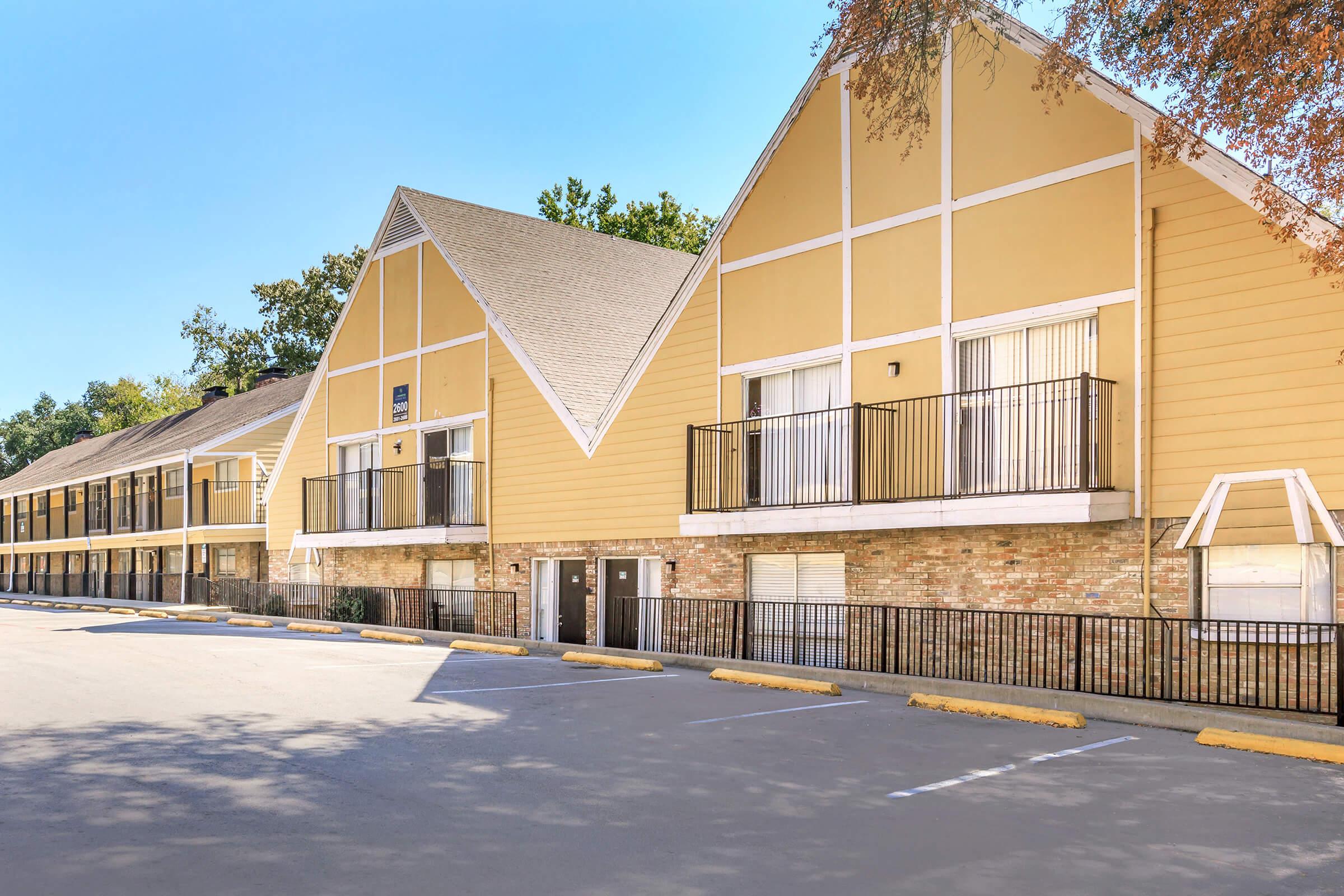 Exterior view of a two-story, yellow apartment building with a pitched roof, featuring multiple balconies. The building has a stone base and is set against a clear blue sky with trees nearby. An empty parking lot is visible in front of the building.