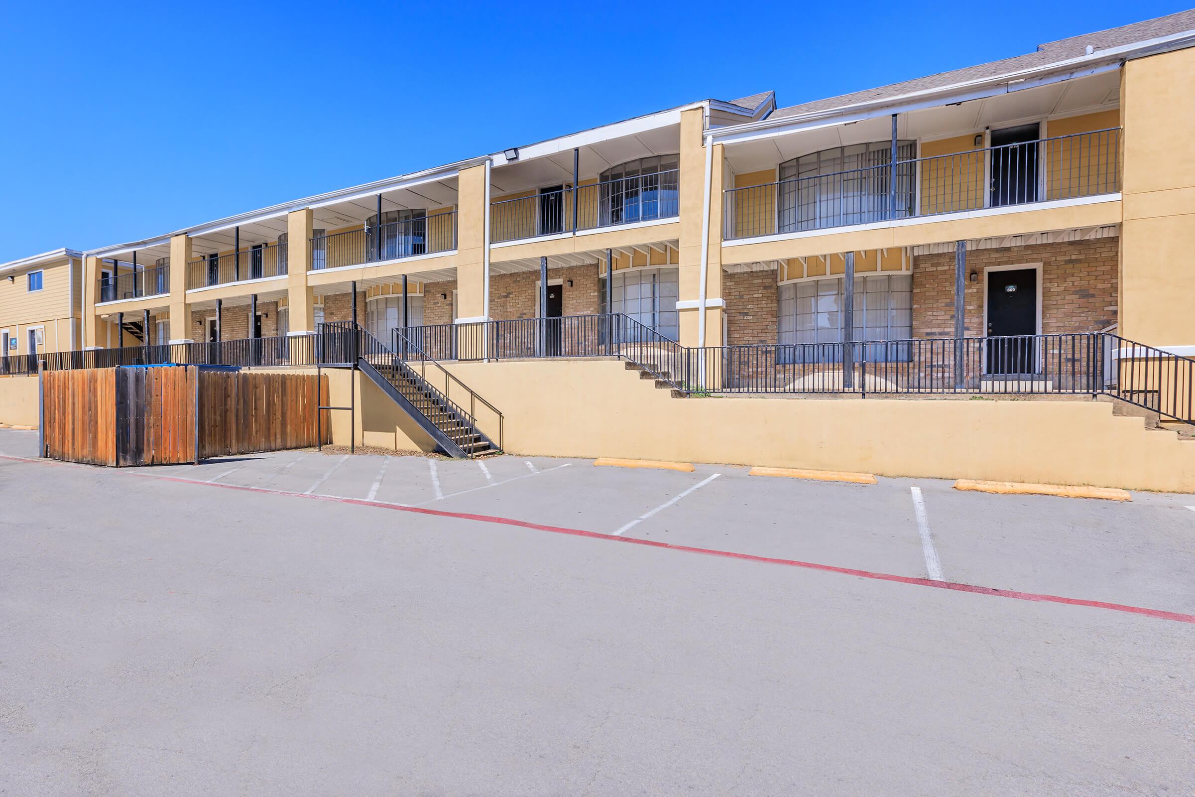A low-rise apartment building featuring multiple units with balconies, non-carpeted outdoor stairs, and a fenced-off parking lot. The exterior is a combination of beige and brick, with a clear blue sky above. Parking spaces are visible in front of the building, which has a well-maintained appearance.