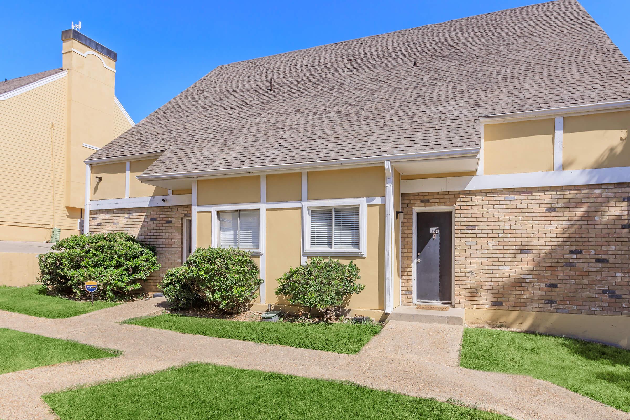 A two-story townhouse with a tan exterior and a sloped roof. The front features a small porch and a door framed by brick walls. Lush green shrubs line the walkway leading up to the entrance, and the area is well-maintained with a grassy lawn under a clear blue sky.