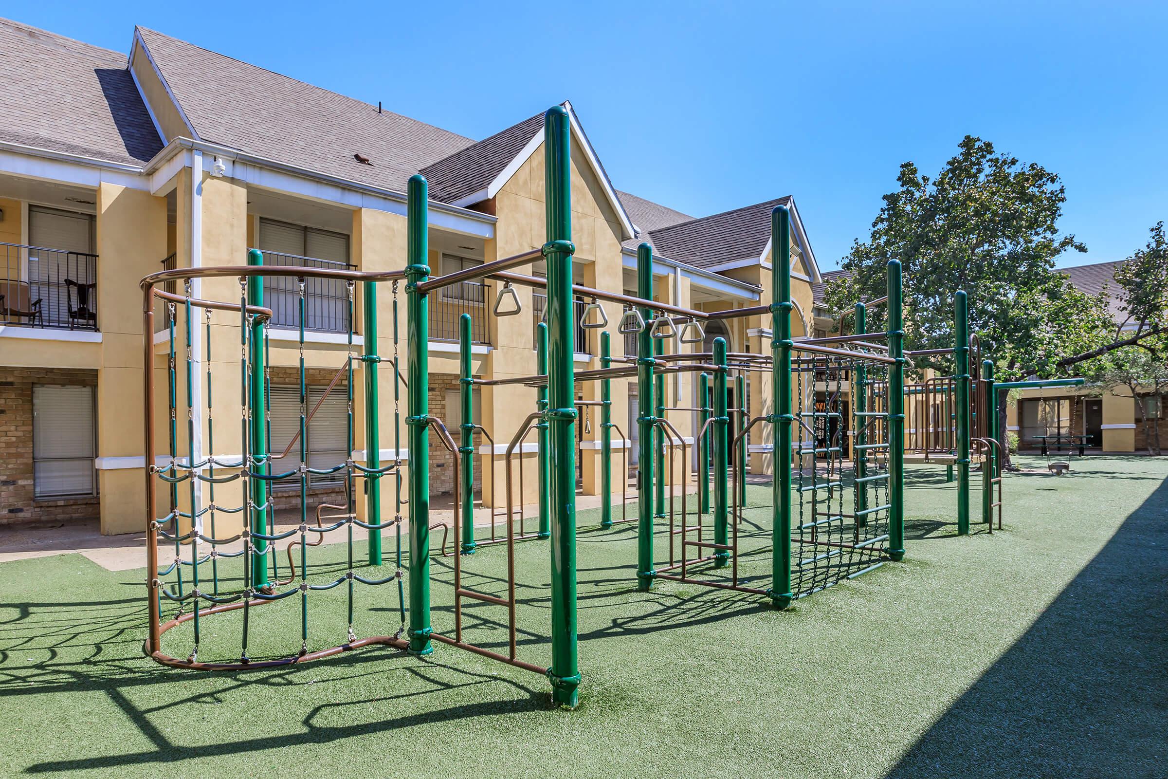 A playground featuring climbing structures and monkey bars set on artificial grass, surrounded by residential buildings. The sky is clear and blue, providing a bright atmosphere for outdoor play.
