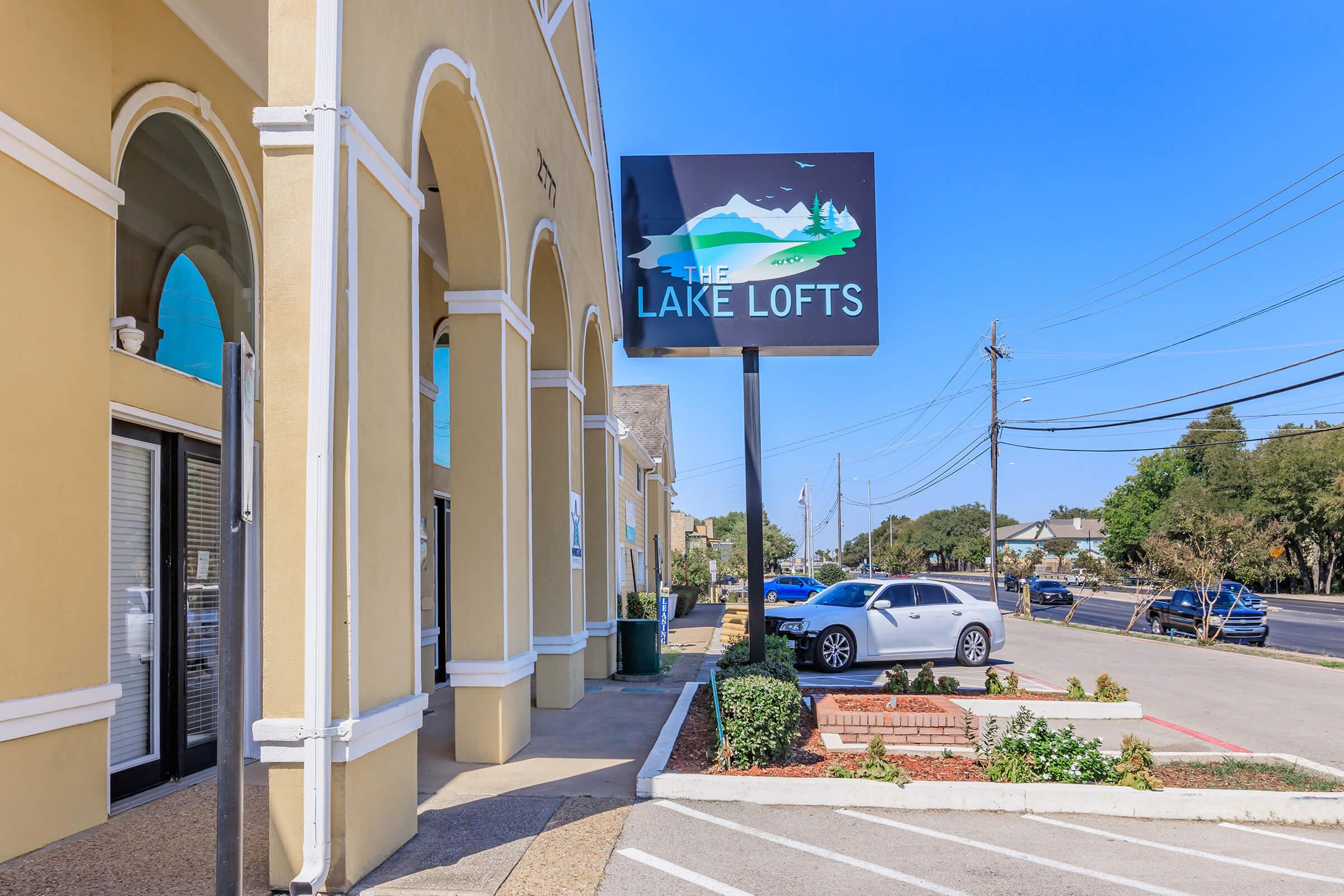Exterior view of The Lake Lofts building featuring a sign with a stylized depiction of a lake and mountains. The entrance has large windows and a door, with a landscaped area in front. The street is visible with parked cars and power lines under a clear blue sky.