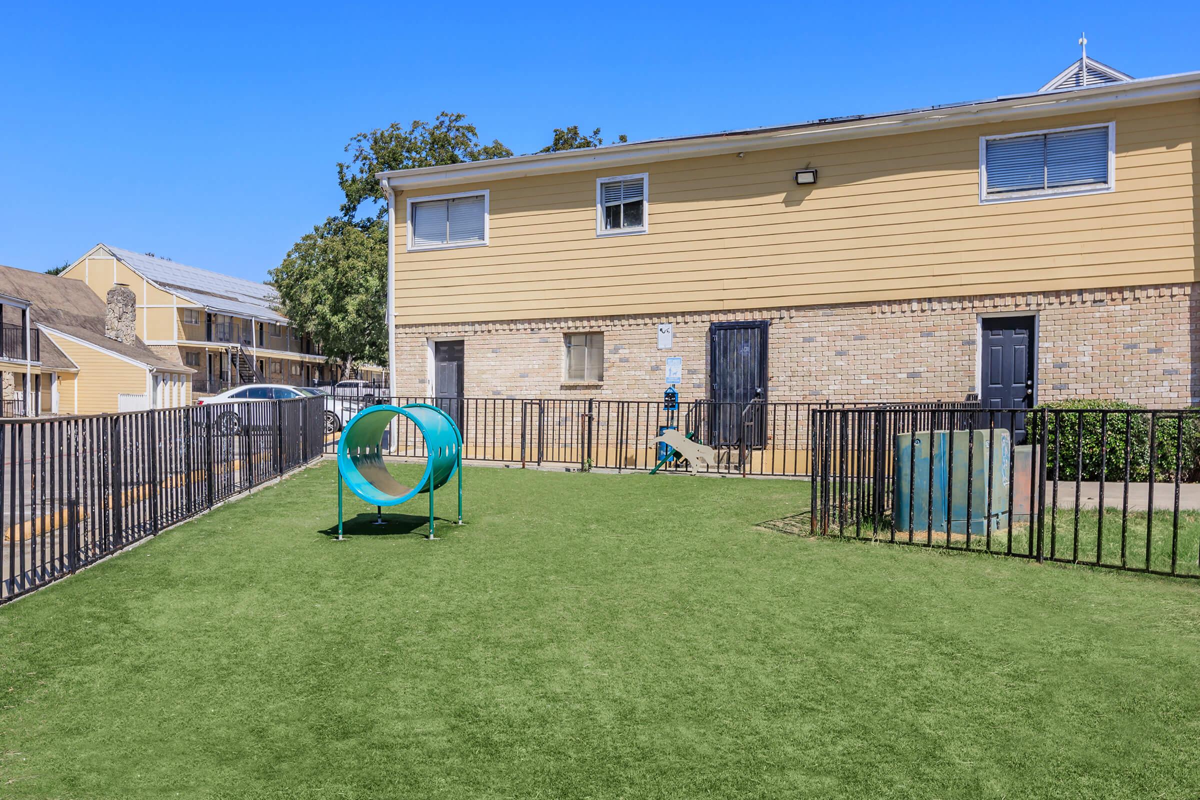 A grassy area with a fenced playground featuring a circular tunnel for children or pets, flanked by two buildings with yellow siding and brick accents. The sky is clear and blue, and there are trees in the background.