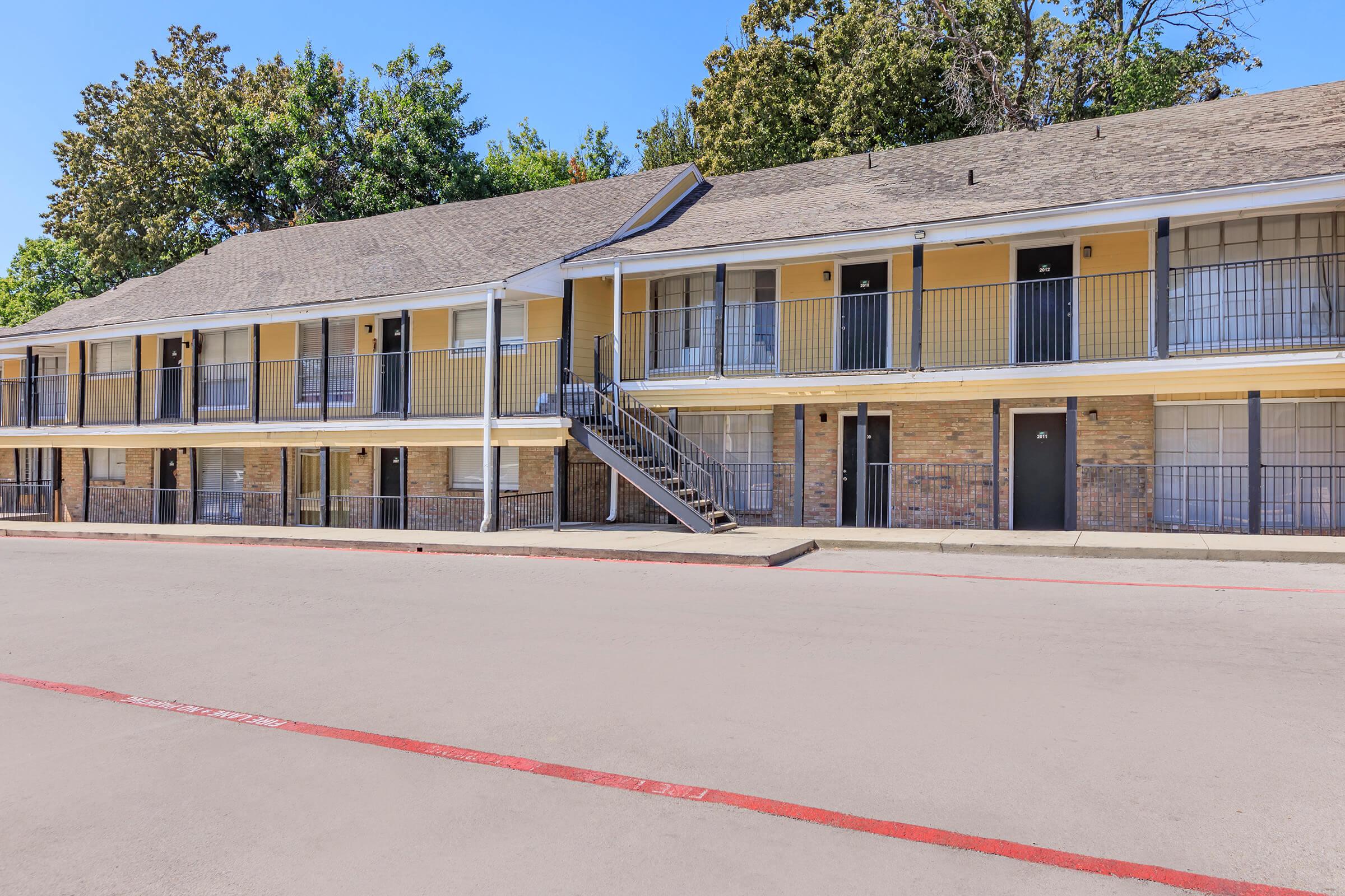 A two-story motel with yellow and brick exterior. The building features multiple rooms with balconies and black railings. A staircase leads to the upper level. The parking lot in front is empty, and trees can be seen in the background under a clear blue sky.