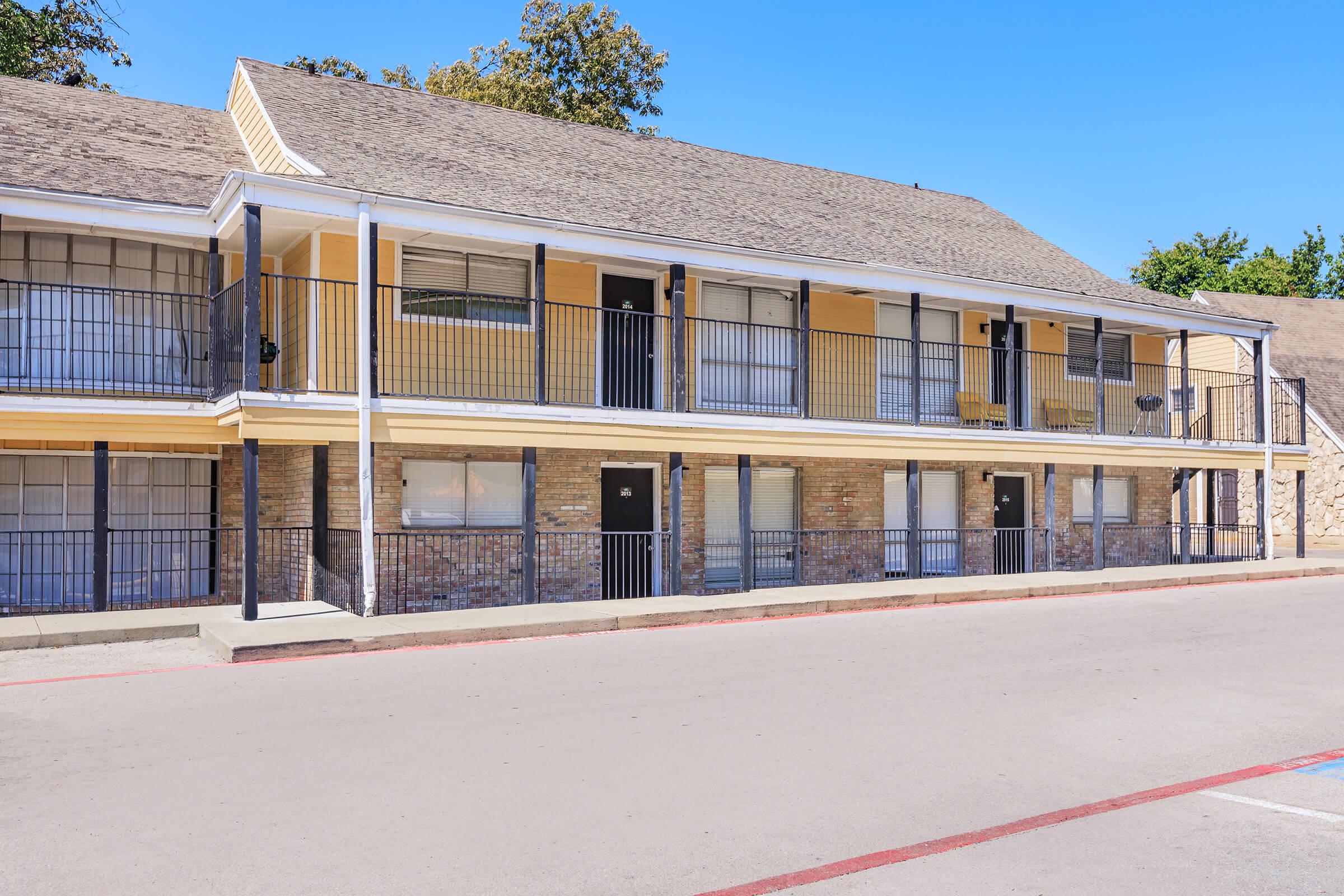 A view of a two-story motel with yellow exterior walls and black railings. The building features multiple rooms with windows, a concrete pathway in front, and parking spaces visible. The sky is clear and blue, with trees in the background.
