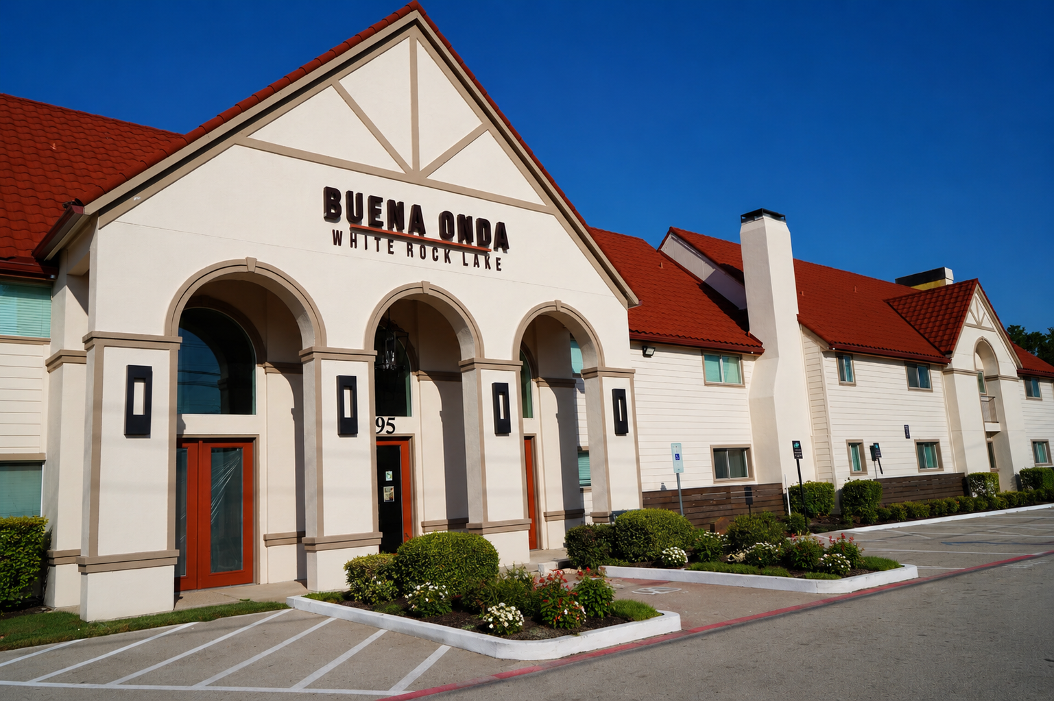 Exterior view of the Buena Onda building at White Rock Lake, featuring a distinct architectural style with arched entrances, large windows, and a red-tiled roof. The surrounding area includes landscaped flower beds and parking spaces, under a clear blue sky.