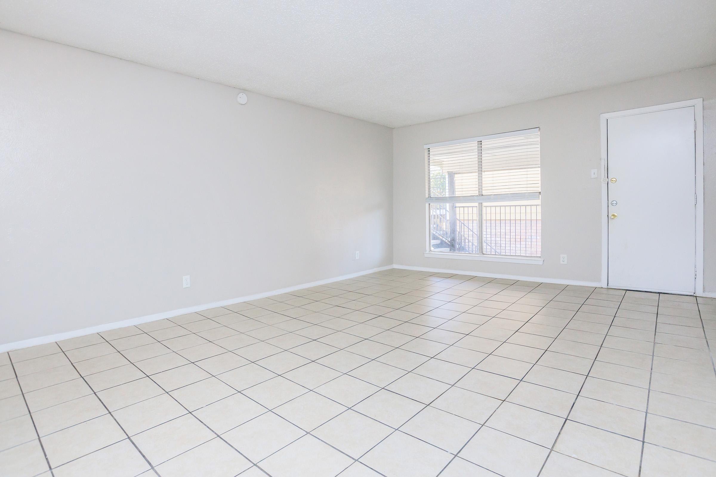 Empty room with light-colored walls and tiled floor. A large window lets in natural light, and there's a door visible on the right side. The space appears clean and uncluttered, ideal for various uses or personal decoration.