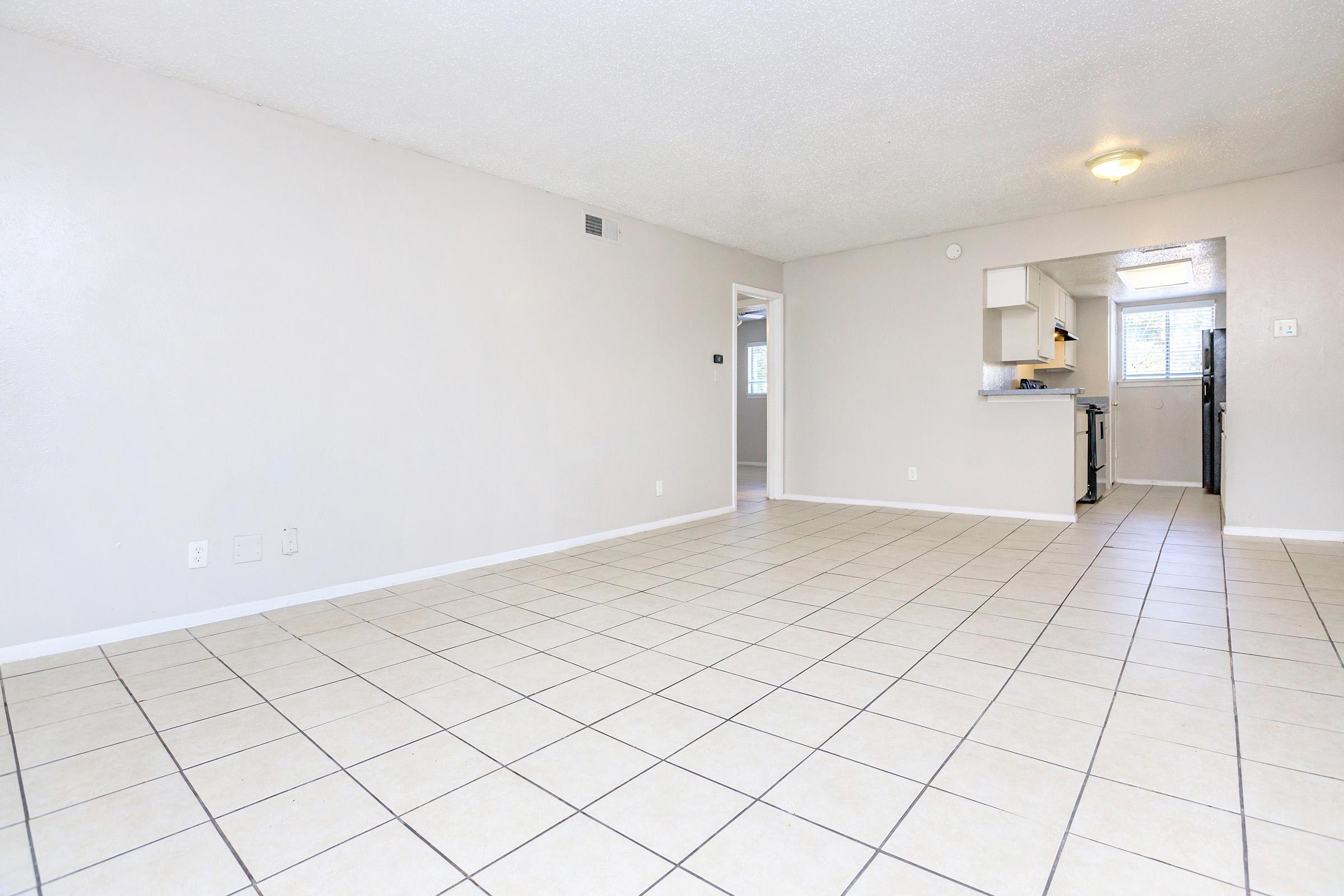Spacious, empty living area with light-colored walls and tiled flooring. Features large windows allowing natural light, with a view towards a kitchen area in the background. The layout is open and inviting, ideal for various furniture arrangements.