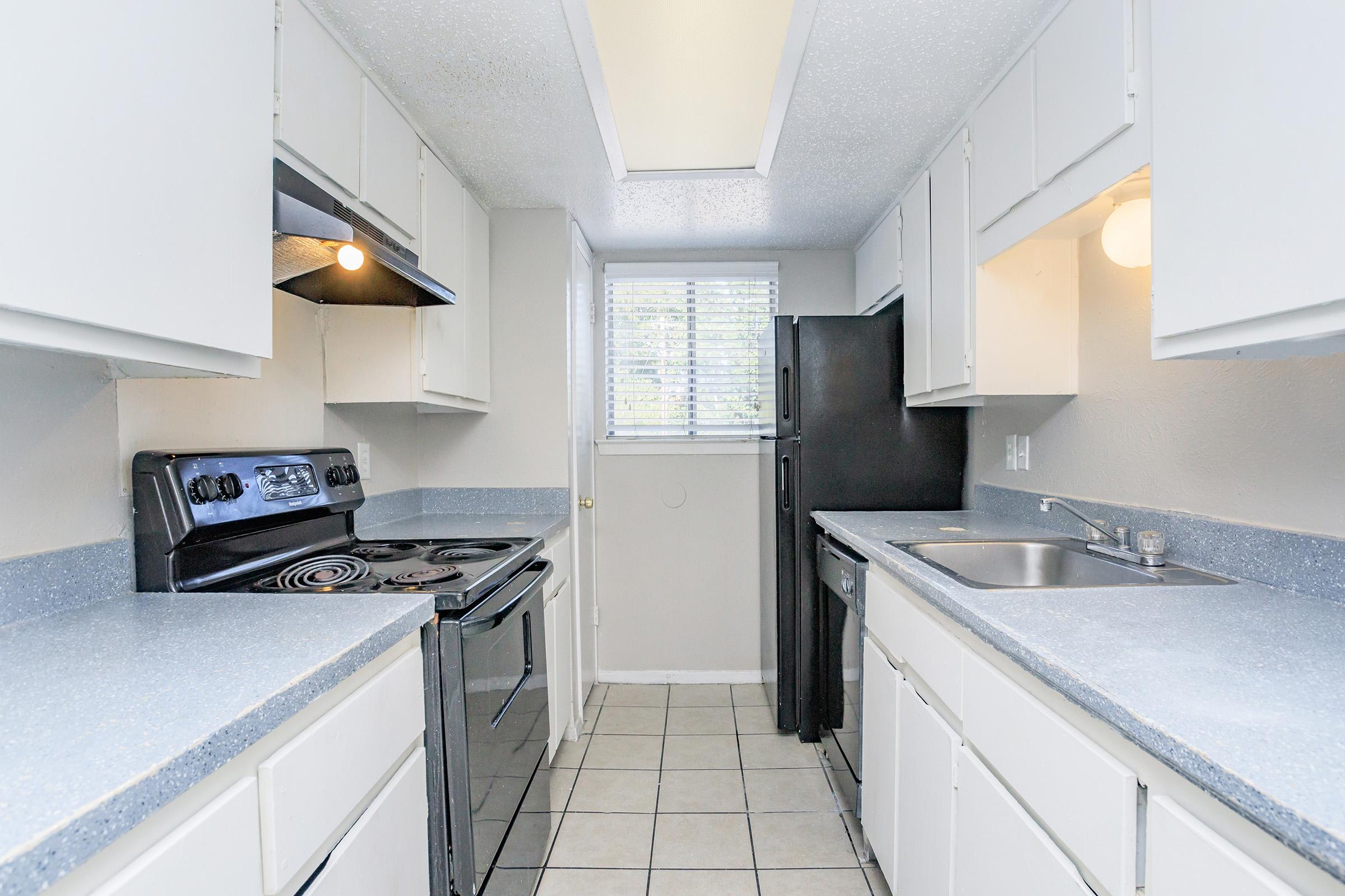 A modern kitchen featuring grey countertops, white cabinets, a black stove and oven, a black refrigerator, and a stainless steel sink. Natural light enters through a window with blinds, illuminating the tile floor.