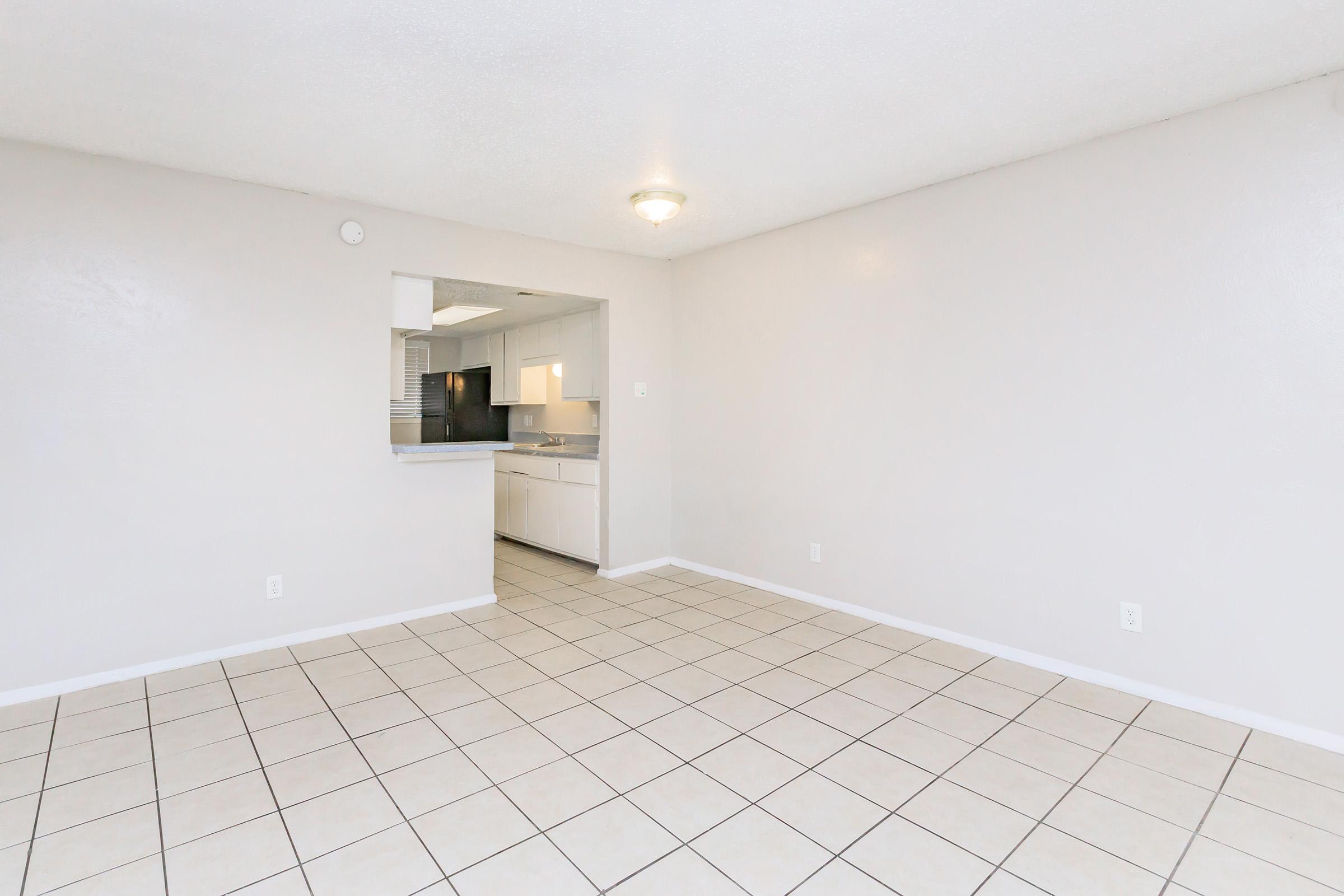 A spacious, empty living area with light-colored walls and tiled flooring. A doorway leads to a kitchen area, which is partially visible and features white cabinets and appliances. Natural light fills the room, creating a bright and open atmosphere.
