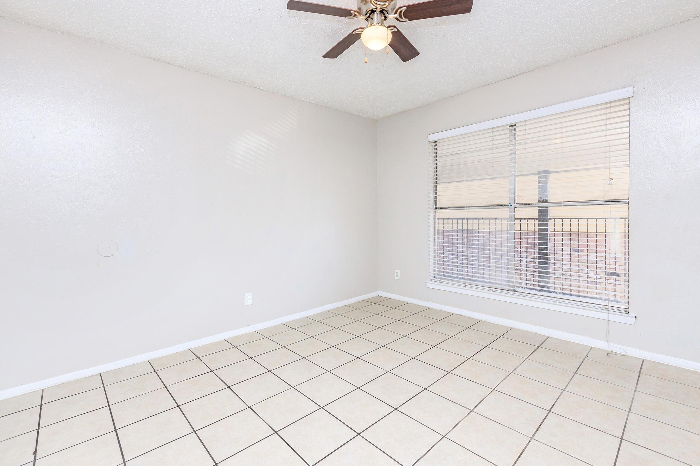 A light-colored room with tiled flooring and a ceiling fan. The walls are bare and the only window features blinds, allowing natural light to enter. The room is empty, creating a spacious and minimalistic appearance.