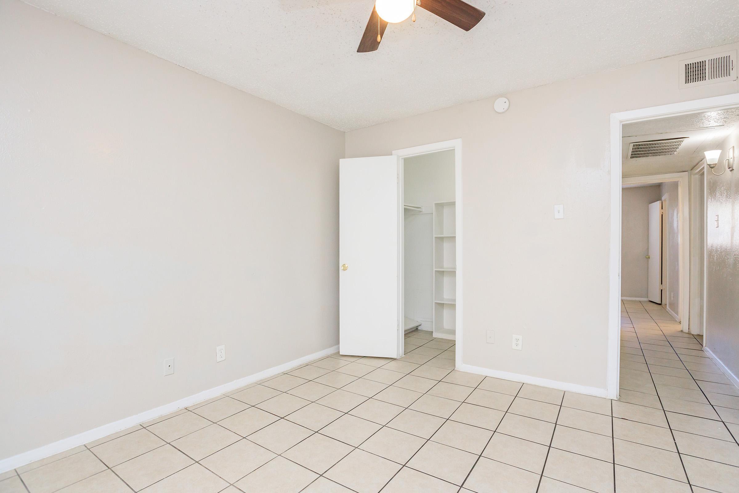 A well-lit room with light-colored walls and tile flooring. There is a ceiling fan, an open closet with shelves, and a doorway leading to another room or hallway. The space appears clean and empty, suitable for various uses.
