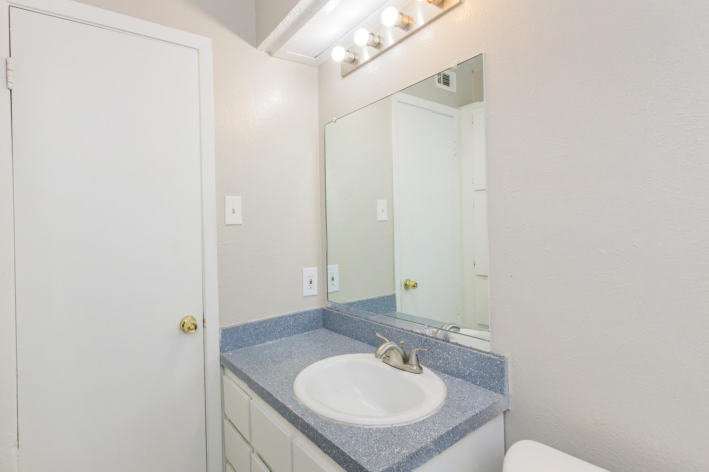 A bathroom featuring a light gray wall, a white sink with a brushed nickel faucet, and a large mirror above the sink. A blue countertop extends beneath the mirror. The room includes a closed door on the left and a glimpse of a cabinet on the right.