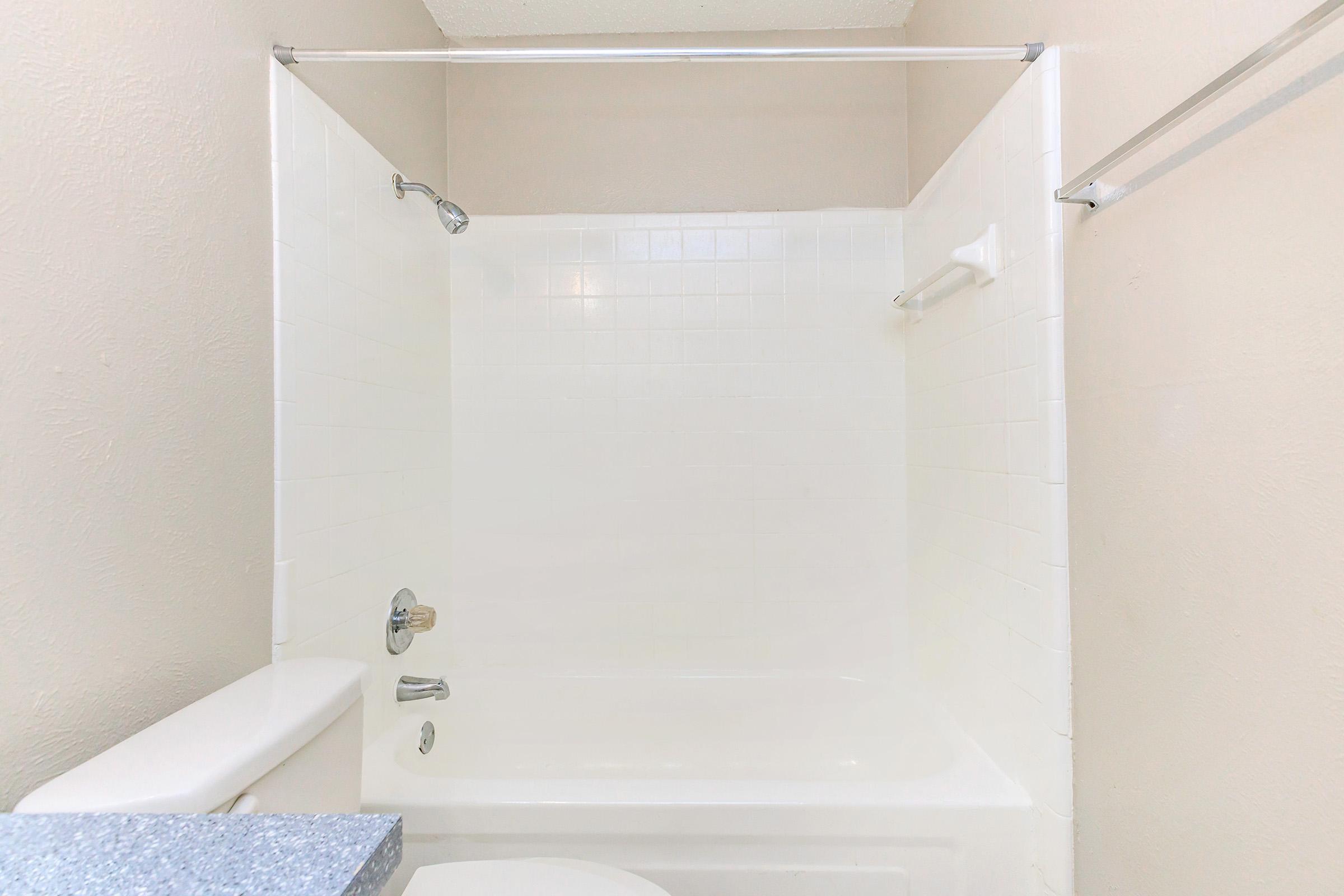 A clean, simple bathroom featuring a white bathtub with a showerhead, white tiled walls, and a large countertop on the left. The overall color scheme is light and neutral, creating a spacious feel.