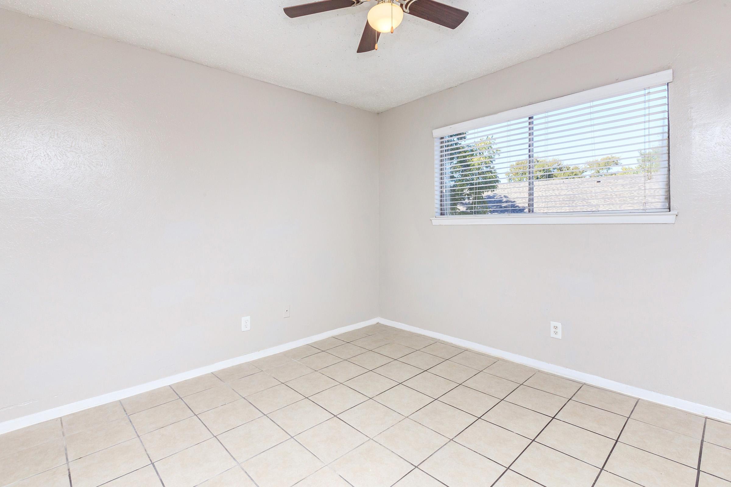 Empty room with light-colored walls and tiled floor. A ceiling fan is installed, and there is a window with blinds allowing natural light to enter. The space appears clean and unfurnished, providing a blank canvas for potential decoration or use.
