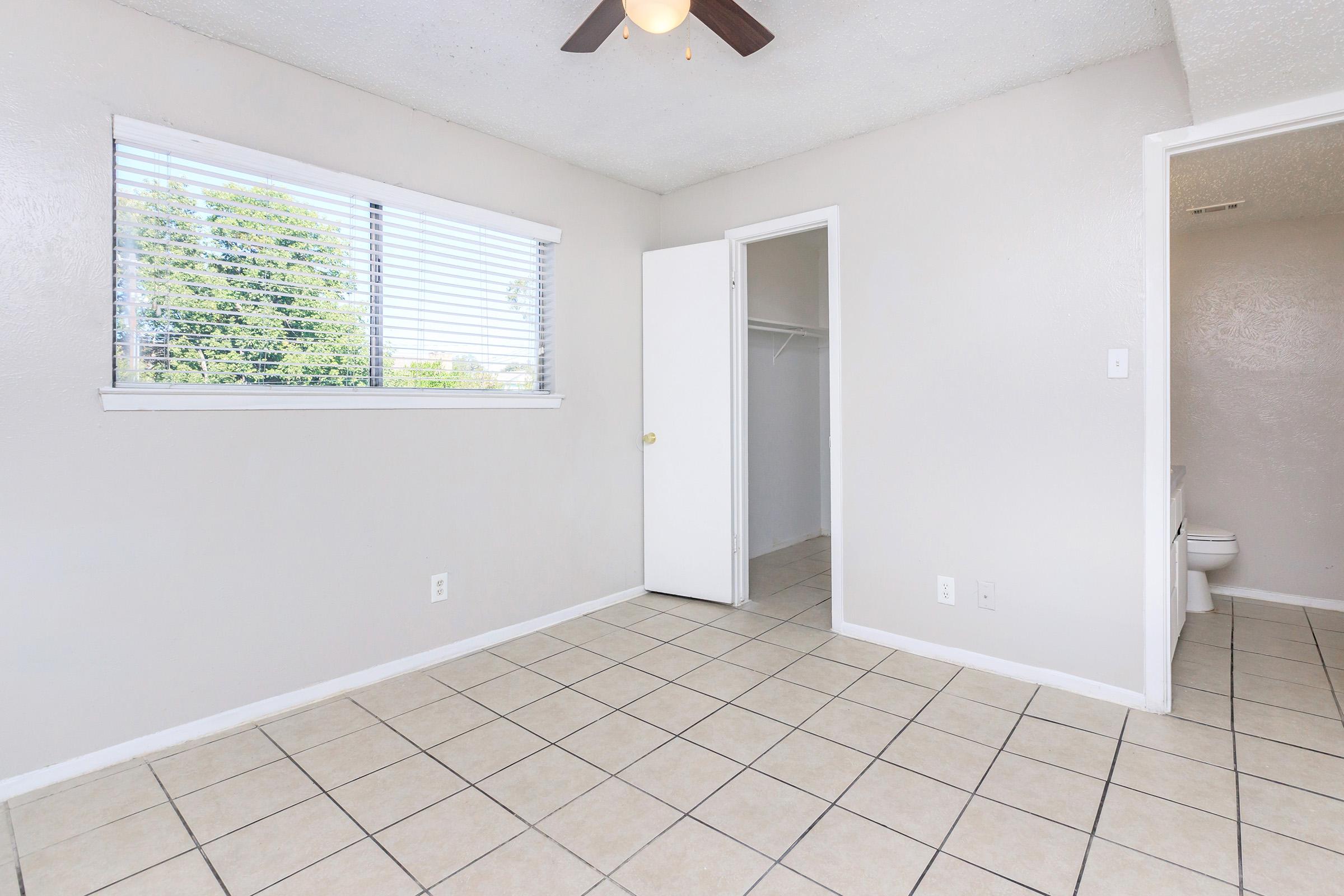 A small, empty room with light-colored walls and tile flooring. It features a window with blinds allowing natural light, a ceiling fan, and a door leading to a closet. There is an adjacent doorway that appears to lead to a bathroom area.