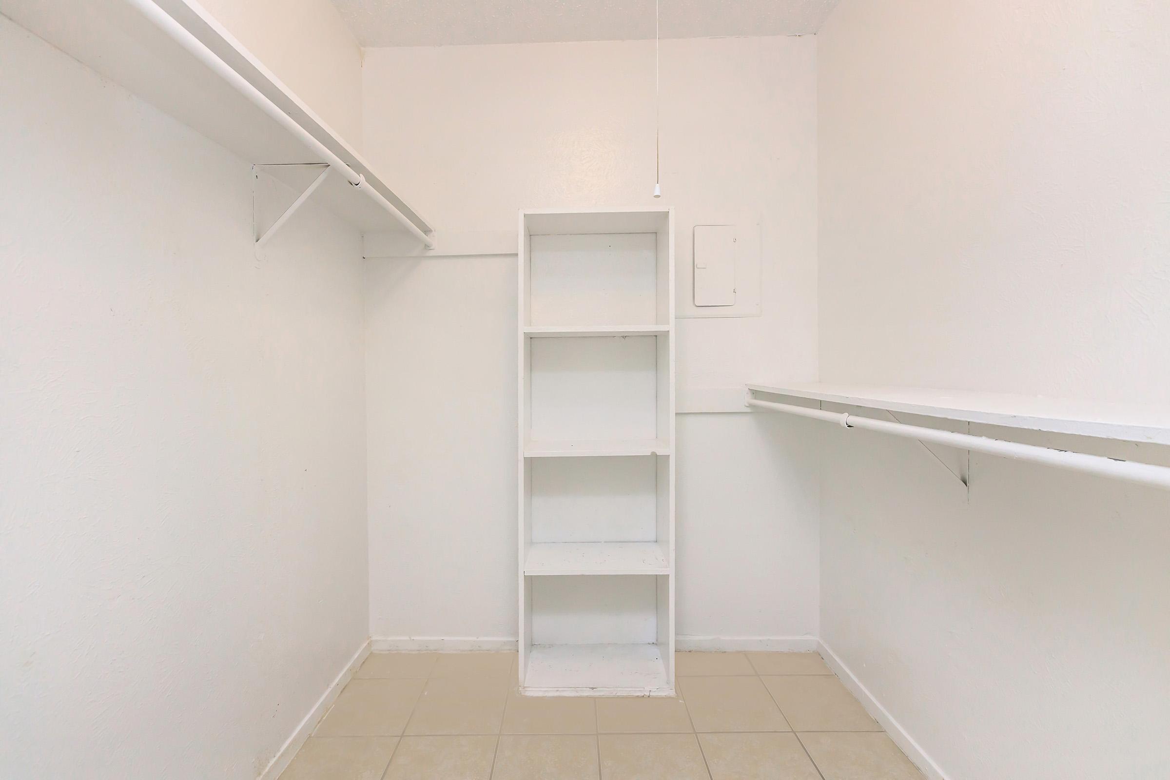 A clean, empty room with white walls and tiled flooring. There is a built-in white shelving unit against one wall, along with two white shelves mounted on the opposite wall. The room is well-lit with natural light and has a minimalistic design, suggesting it could be used as a storage space or closet.