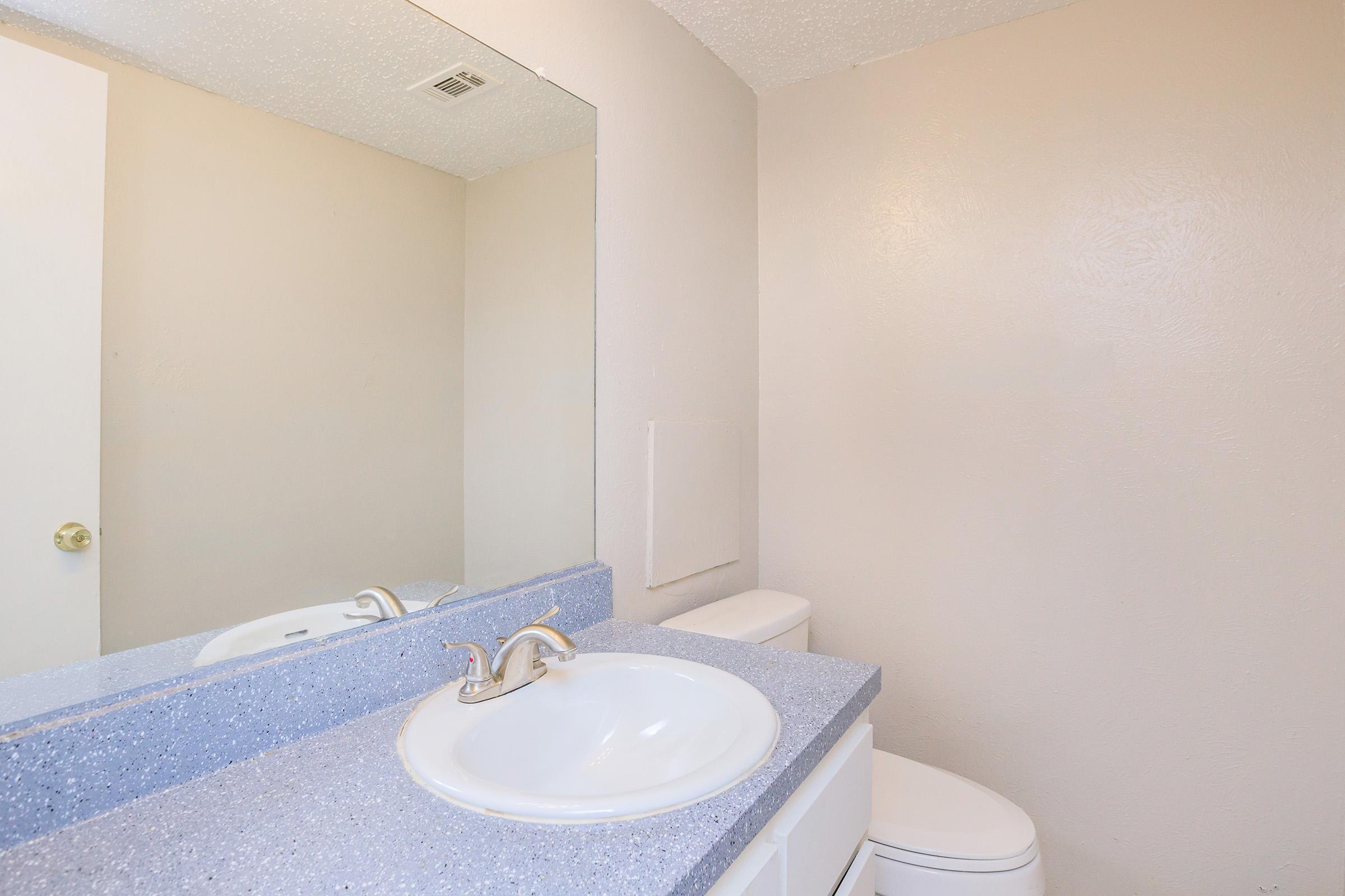A clean, simple bathroom featuring a light-colored countertop with a built-in sink, a large mirror above, and a white toilet. The walls are painted a soft beige, creating a neutral and airy atmosphere. The flooring is not visible in the image.