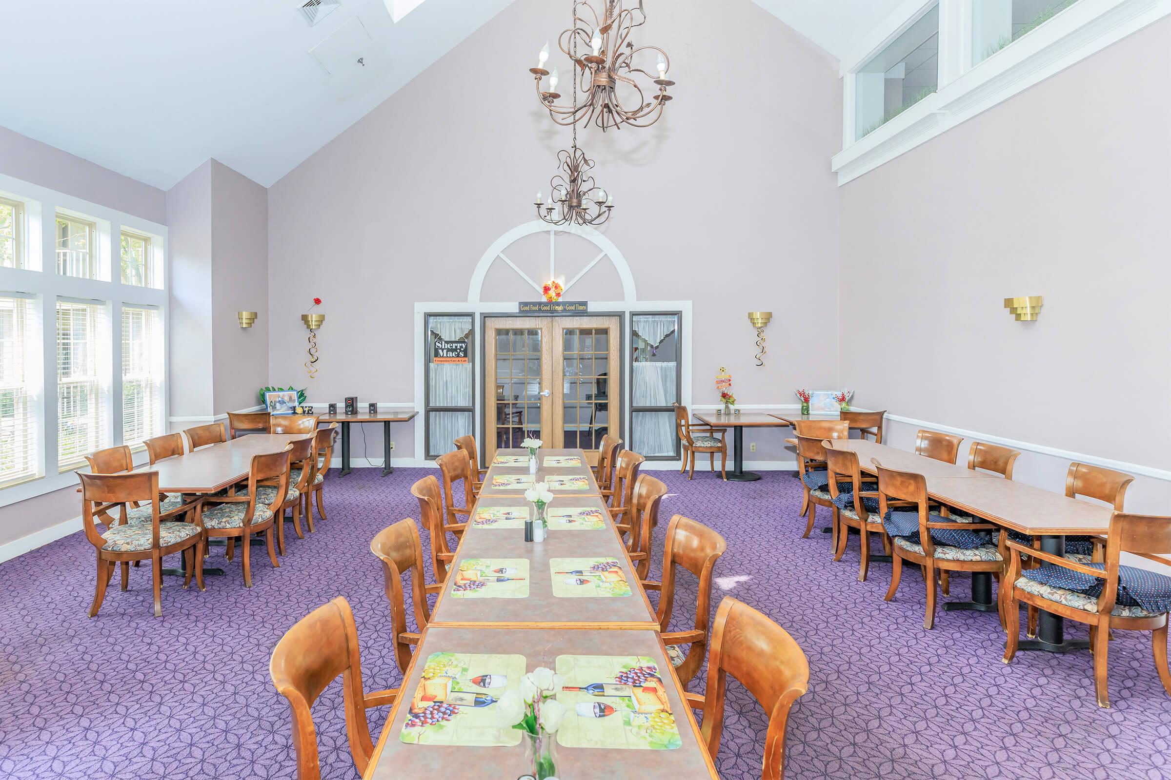 A spacious dining area featuring numerous wooden tables and chairs arranged in two long rows. The floor is covered with a purple carpet, and the walls are painted a soft light color. Natural light enters through large windows, and decorative elements, including a chandelier and wall accents, enhance the inviting atmosphere.