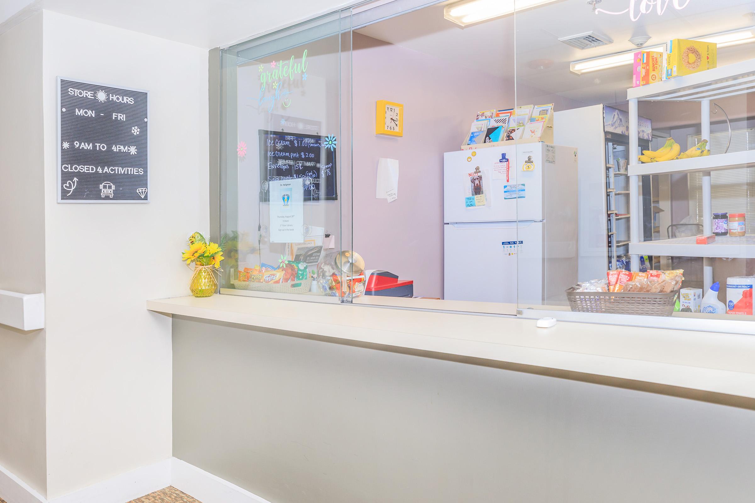 A bright, welcoming reception area with a service counter. A menu board displays store hours. Behind the counter, a refrigerator and shelves are stocked with food items. Fresh flowers add a cheerful touch, enhancing the inviting atmosphere of the space.