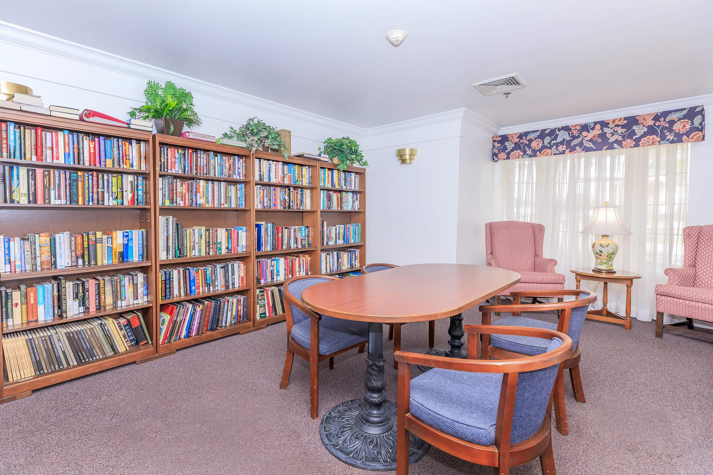 A cozy reading nook featuring a wooden table surrounded by four chairs. In the background, there is a tall bookshelf filled with a variety of books. To the side, two comfortable pink upholstered chairs and a lamp illuminate the space, while a floral-patterned curtain adds a decorative touch to the window.