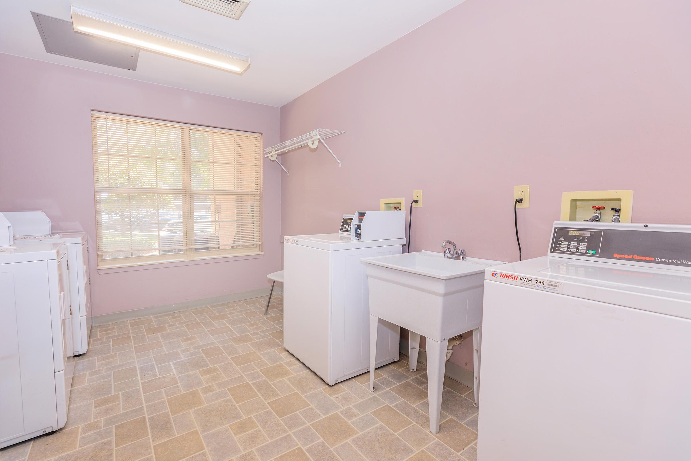 A clean laundry room featuring white washing machines and a sink, with a light pink wall and beige tiled floor. Natural light filters in through a window covered with blinds, creating a bright and inviting atmosphere.