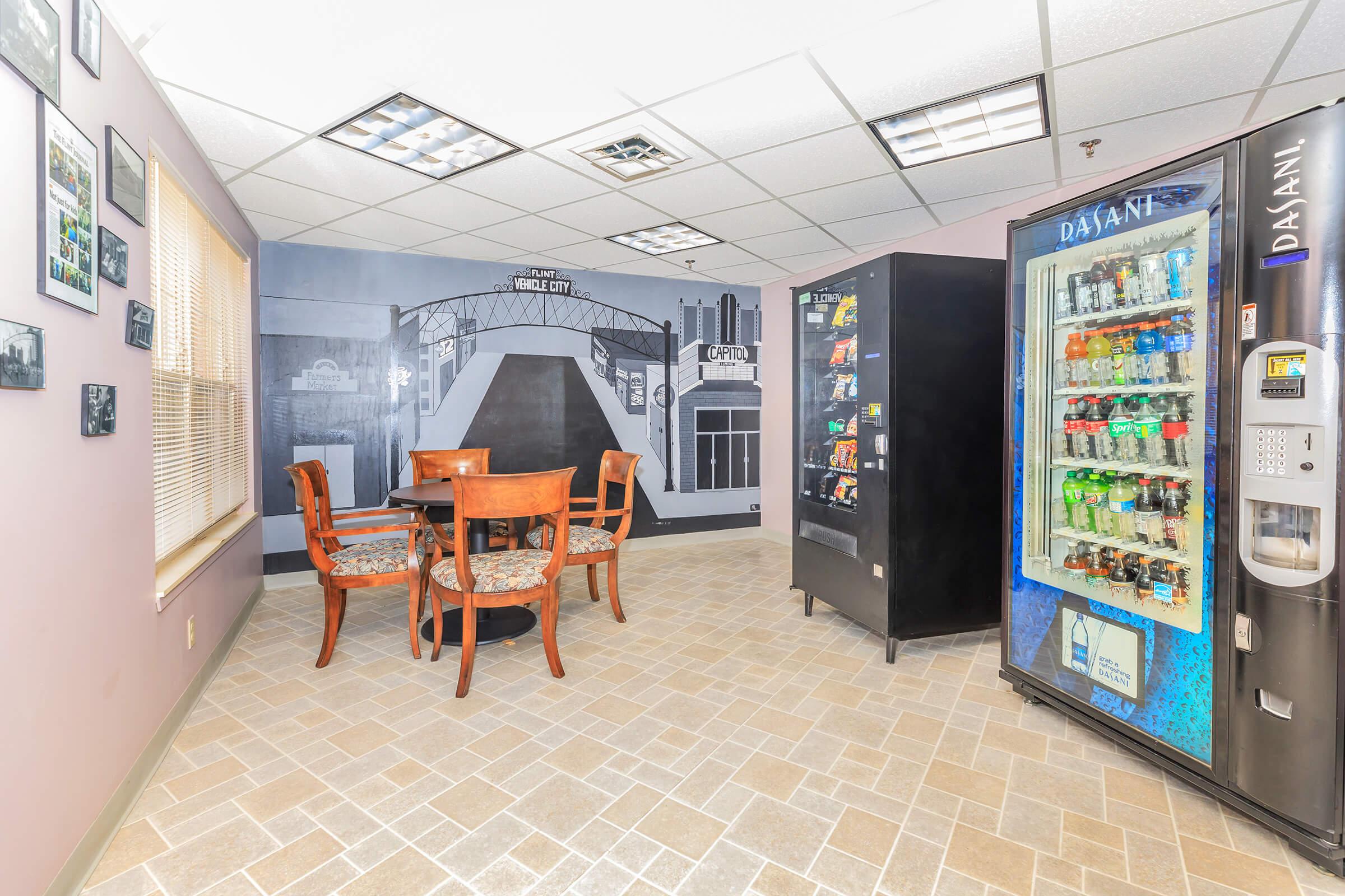 A break room with a table and four chairs, featuring a vending machine stocked with snacks and drinks. The wall has a mural depicting a cityscape. Natural light enters through a window, and the floor is tiled with light-colored squares.