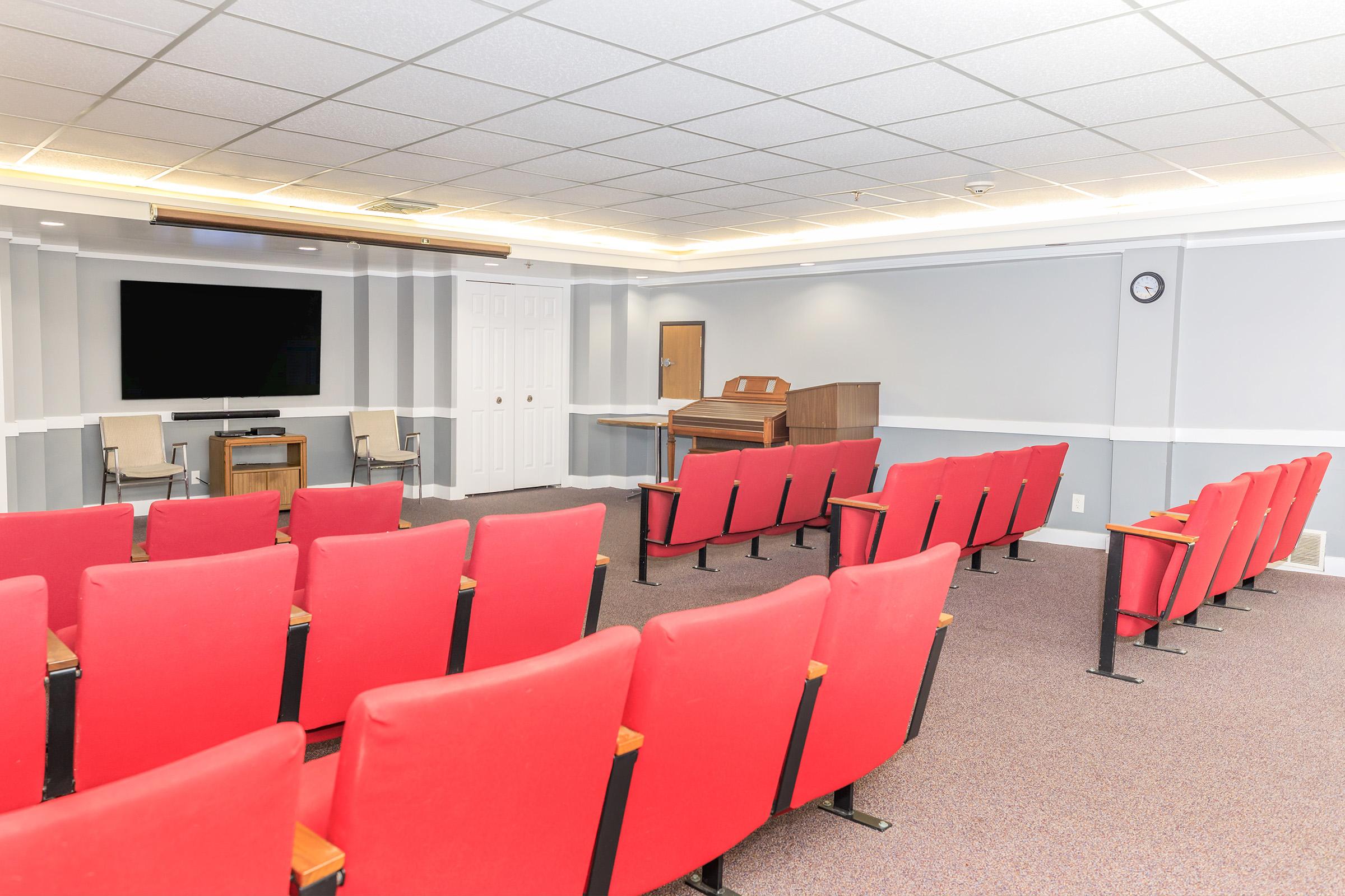 A bright, well-lit room featuring red theater-style seating arranged in rows, facing a large screen at the front. In the background, there's a wooden piano and a clock on the wall. The room has a clean and modern design, suitable for presentations or performances.