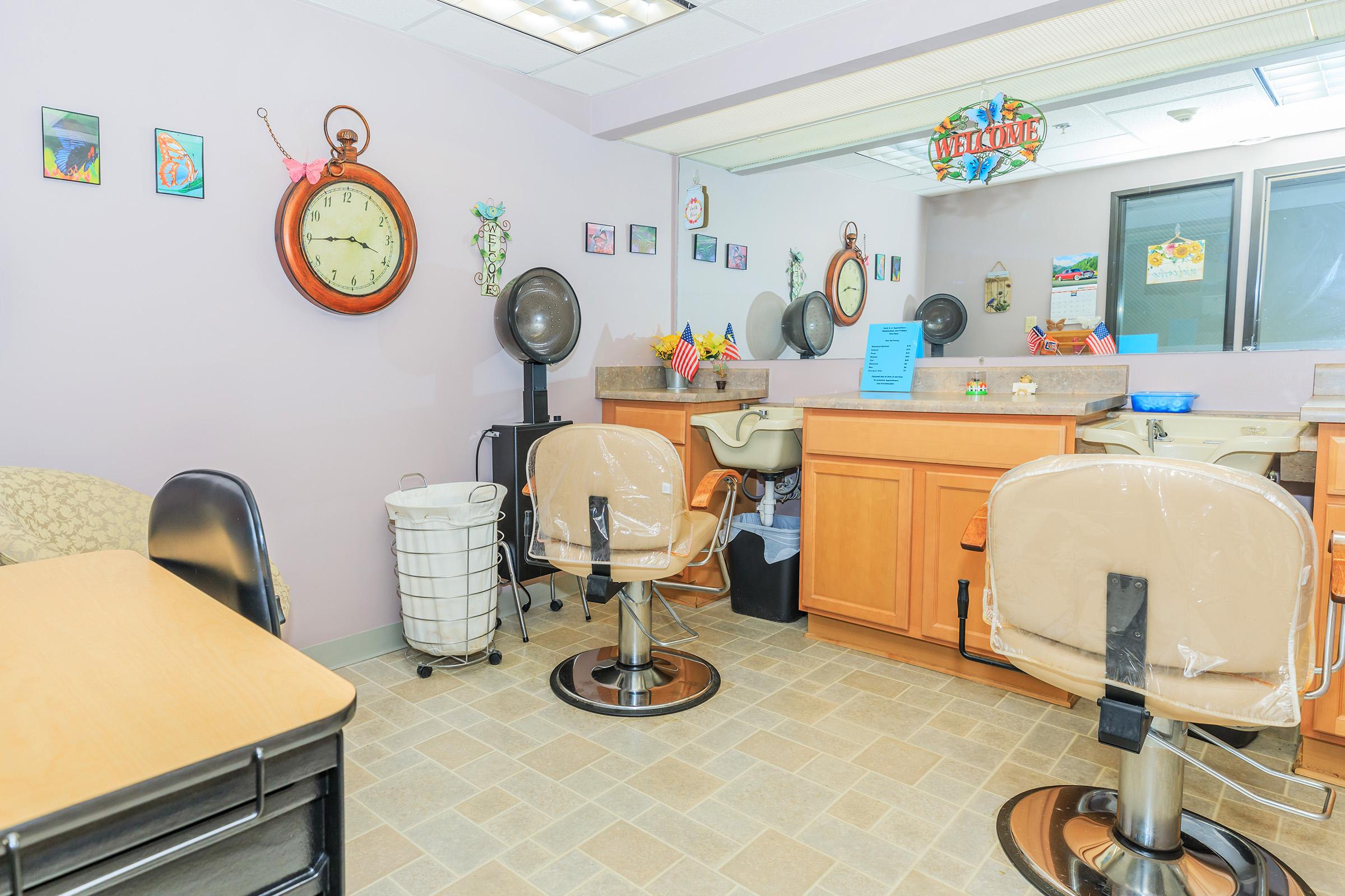 A brightly lit salon interior featuring two styling chairs with plastic covers, a mirror, and a counter with salon supplies. The walls are adorned with colorful artwork and there is a clock and various decorative items. A laundry basket is visible in the corner.