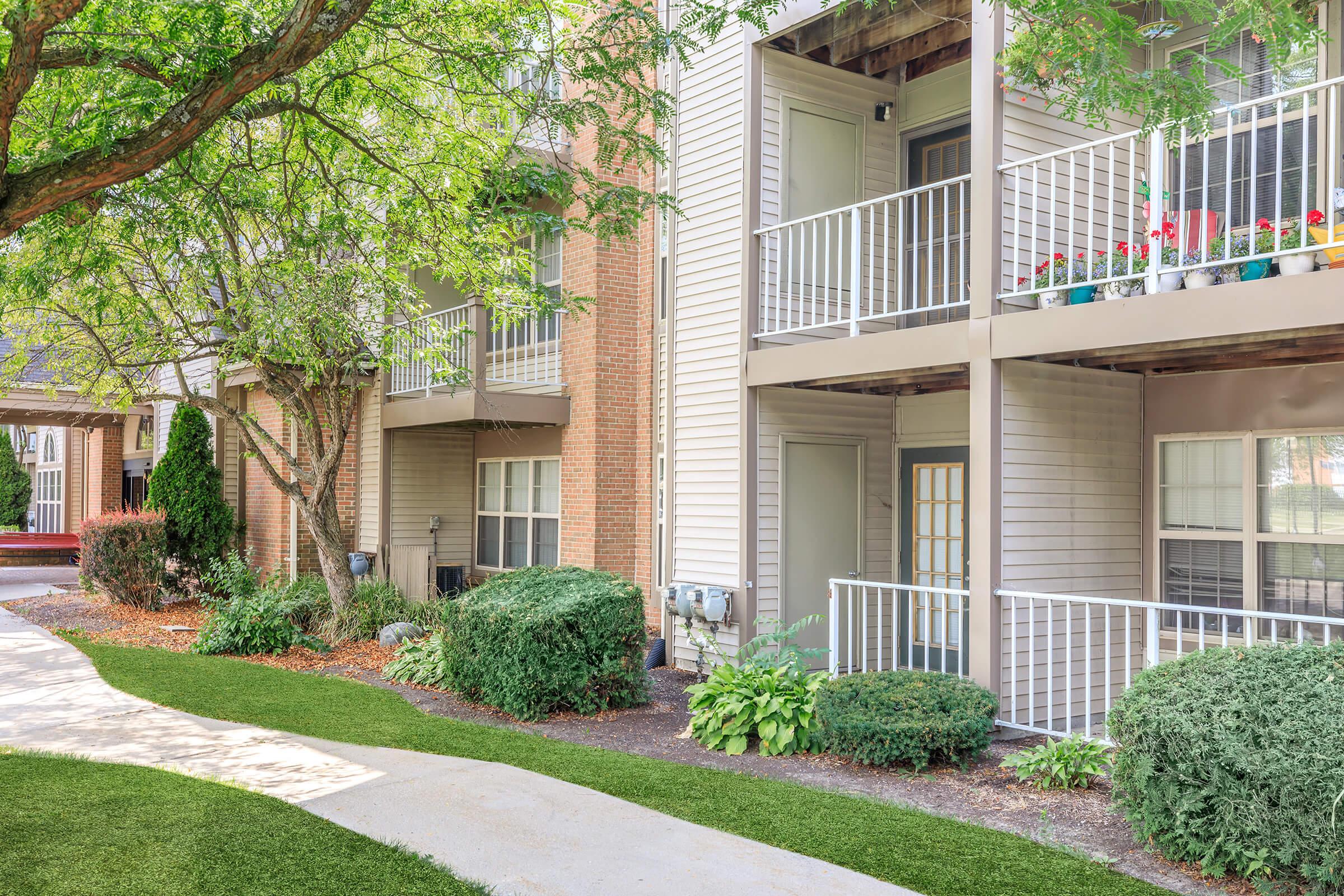 Two apartment buildings with balconies adorned with potted plants. The pathway in front is lined with green grass and shrubs. Trees provide shade, creating a pleasant outdoor environment. The scene is well-maintained, indicative of a residential community.