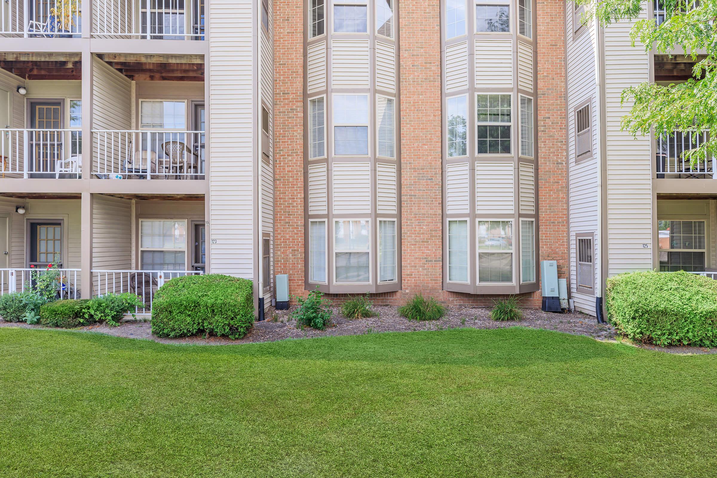 A view of a three-story residential building with balconies and landscaped green grass in the foreground. The building features light-colored siding, with brick accents and lush shrubs framing the lower level. No people are visible; the area appears well-maintained and inviting.