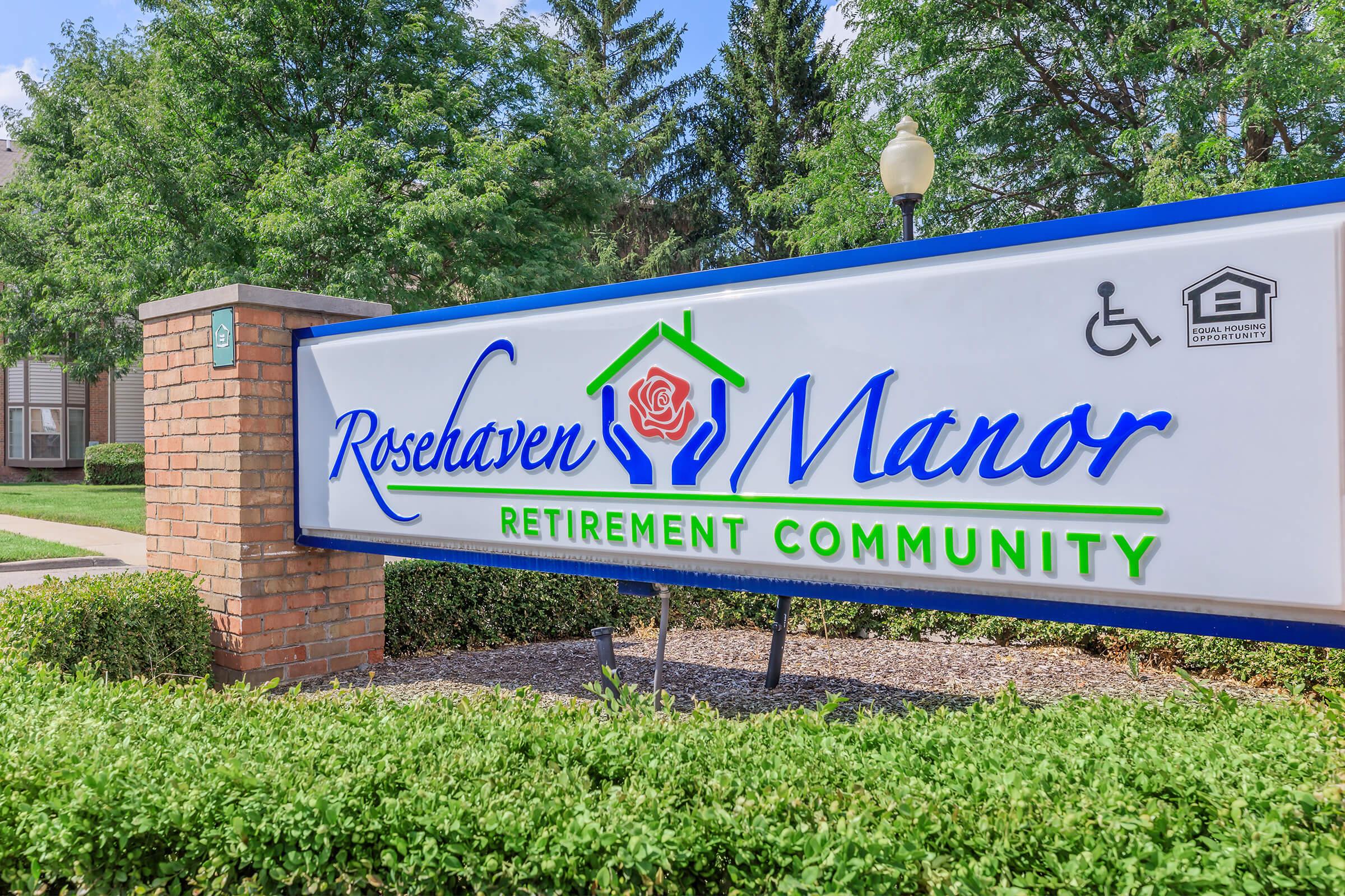 Sign for Rosehaven Manor Retirement Community, featuring a green house logo with a rose. The sign indicates amenities for accessibility and includes trees and landscaping in the background.
