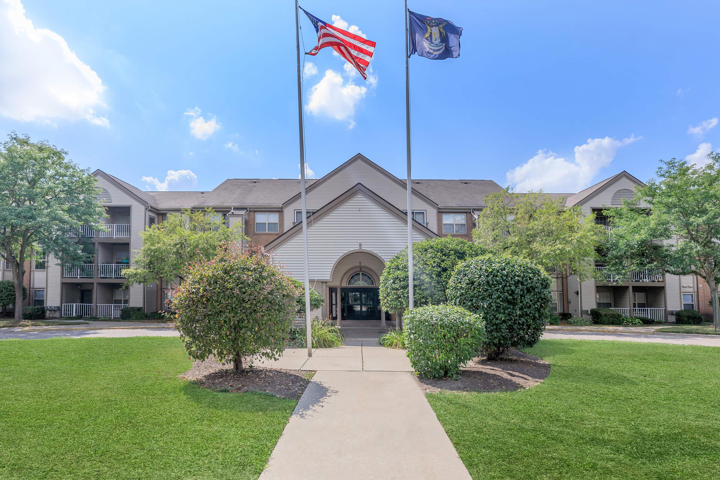 A entrance view of an apartment complex featuring well-maintained landscaping, including shrubs and grass, with an American flag and a state flag flying in front of the building. The sky is clear and blue with a few clouds visible.