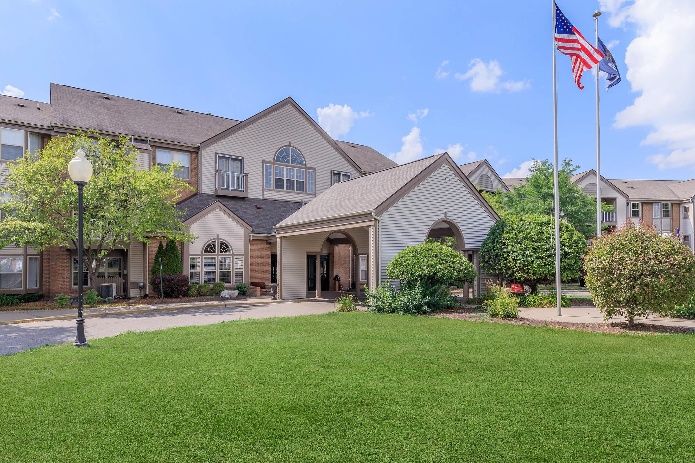 A well-maintained residential building with a welcoming entrance, surrounded by lush green lawns and well-kept shrubbery. The flagpole displays the American flag and another state flag, indicating a sense of community. Bright blue sky with scattered clouds enhances the inviting atmosphere.
