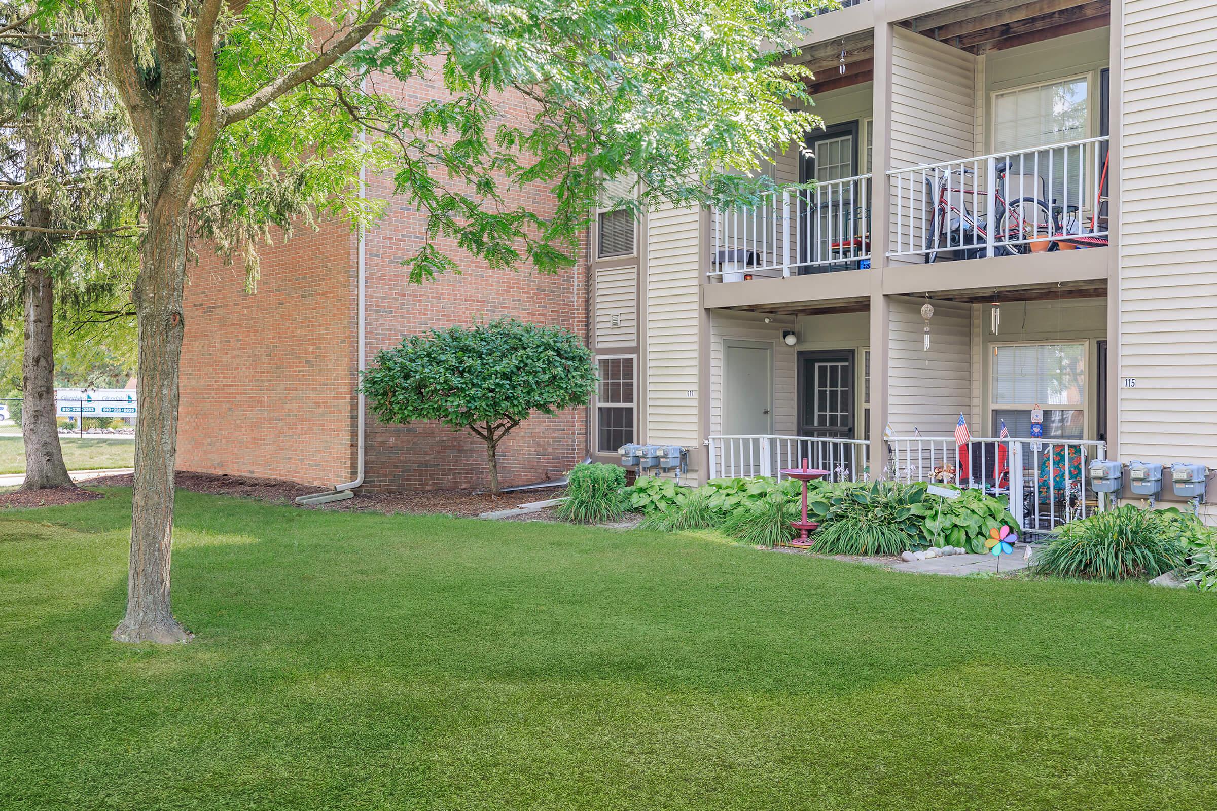 A well-maintained lawn in front of a residential building, featuring a patio with chairs and potted plants. Nearby, a small bush adds greenery, and the exterior includes a mix of light-colored siding and a brick wall. The setting is peaceful, with trees providing shade.