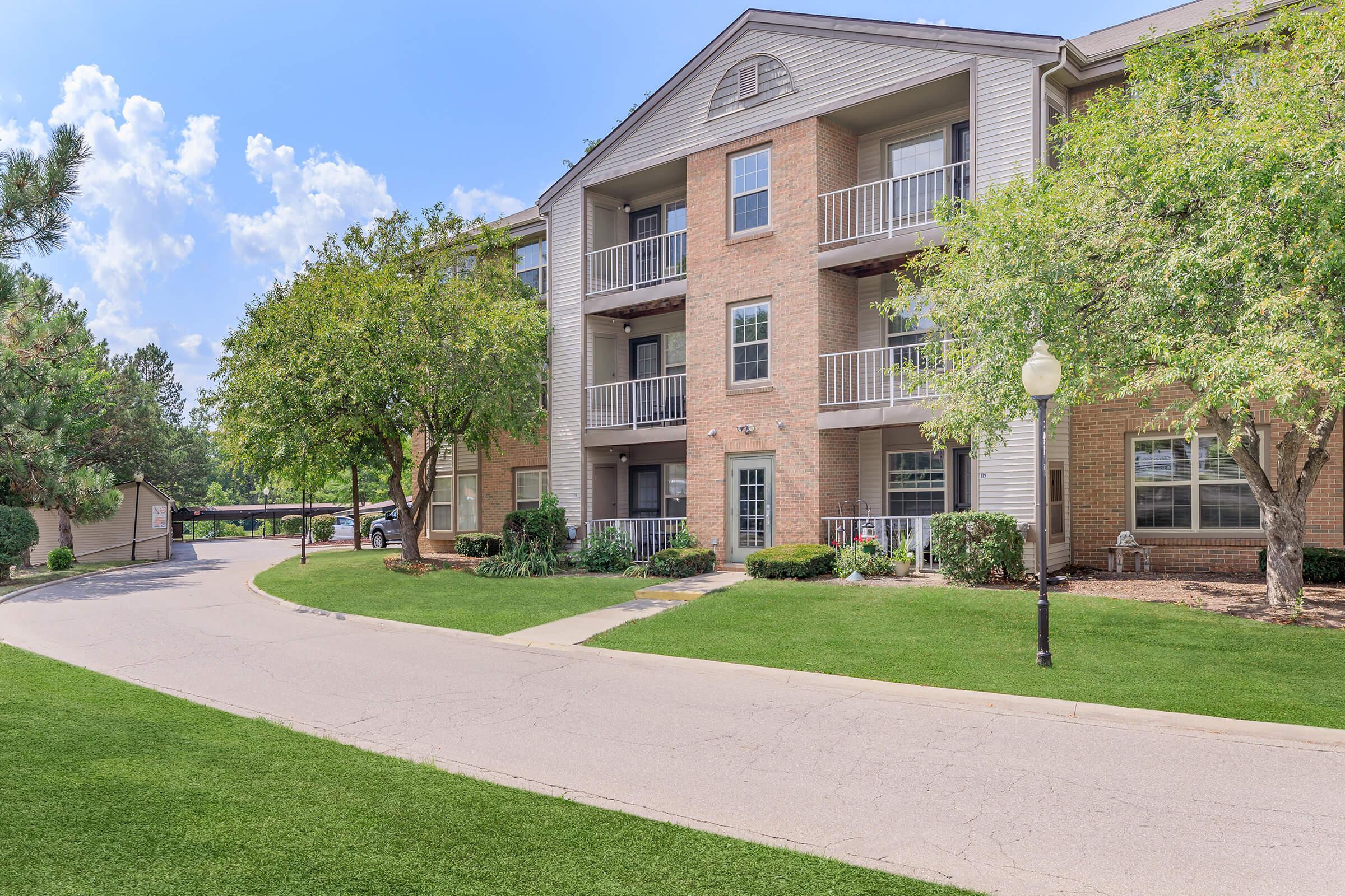 A modern three-story apartment building with a mix of brick and siding, surrounded by green lawns and trees. The pathway leads alongside the building, with balconies visible on each floor. A carport is seen in the background, under a partly cloudy blue sky.