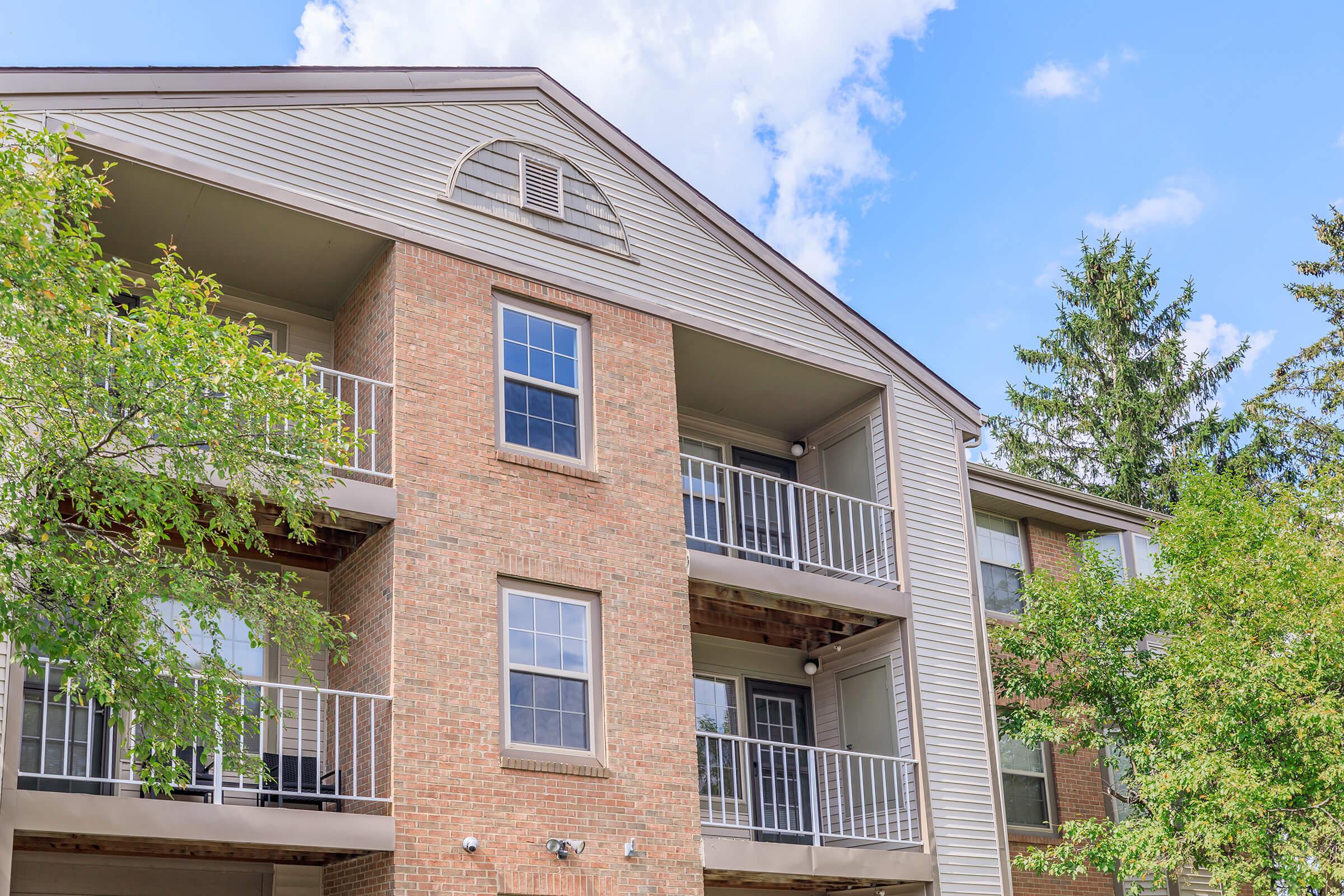 Multi-story residential building with a brick exterior and gray siding. The structure features balconies, large windows, and lush green trees surrounding it under a partly cloudy sky.