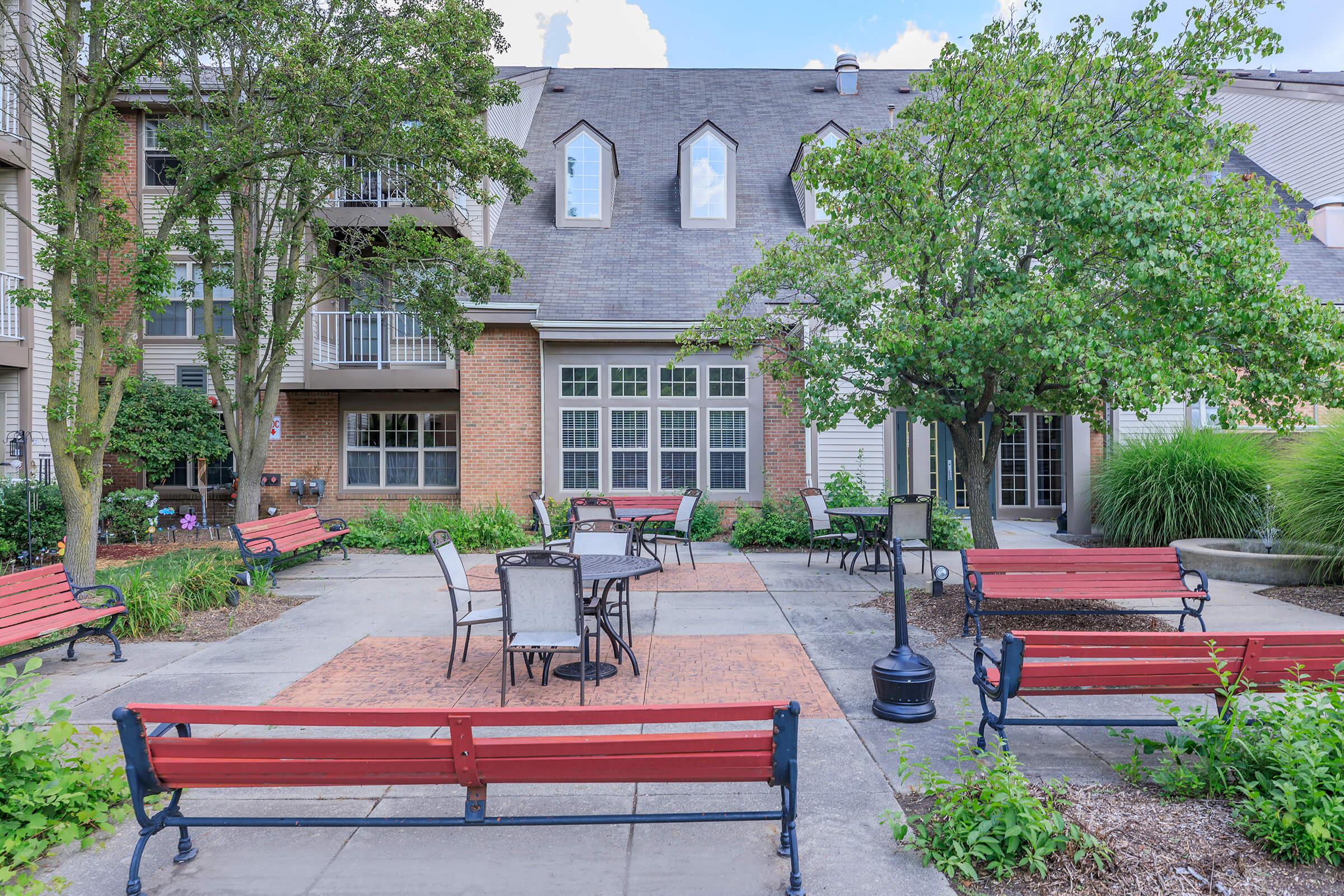 A peaceful courtyard featuring several red benches and metal tables surrounded by greenery and flowering plants. The background includes a building with multiple windows and a distinctive roof design, creating a serene outdoor space for relaxation or gathering.