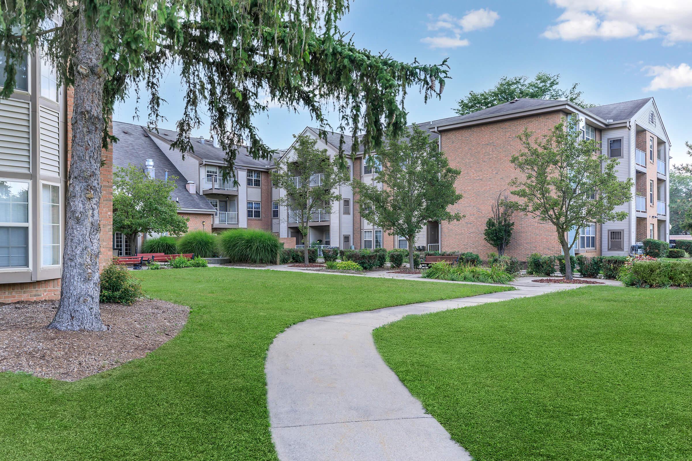 A landscaped courtyard featuring well-maintained greenery, trees, and shrubs, with a curved walkway leading through the area. Surrounding buildings are residential, showcasing a mix of brick and modern architecture, providing a serene outdoor setting.
