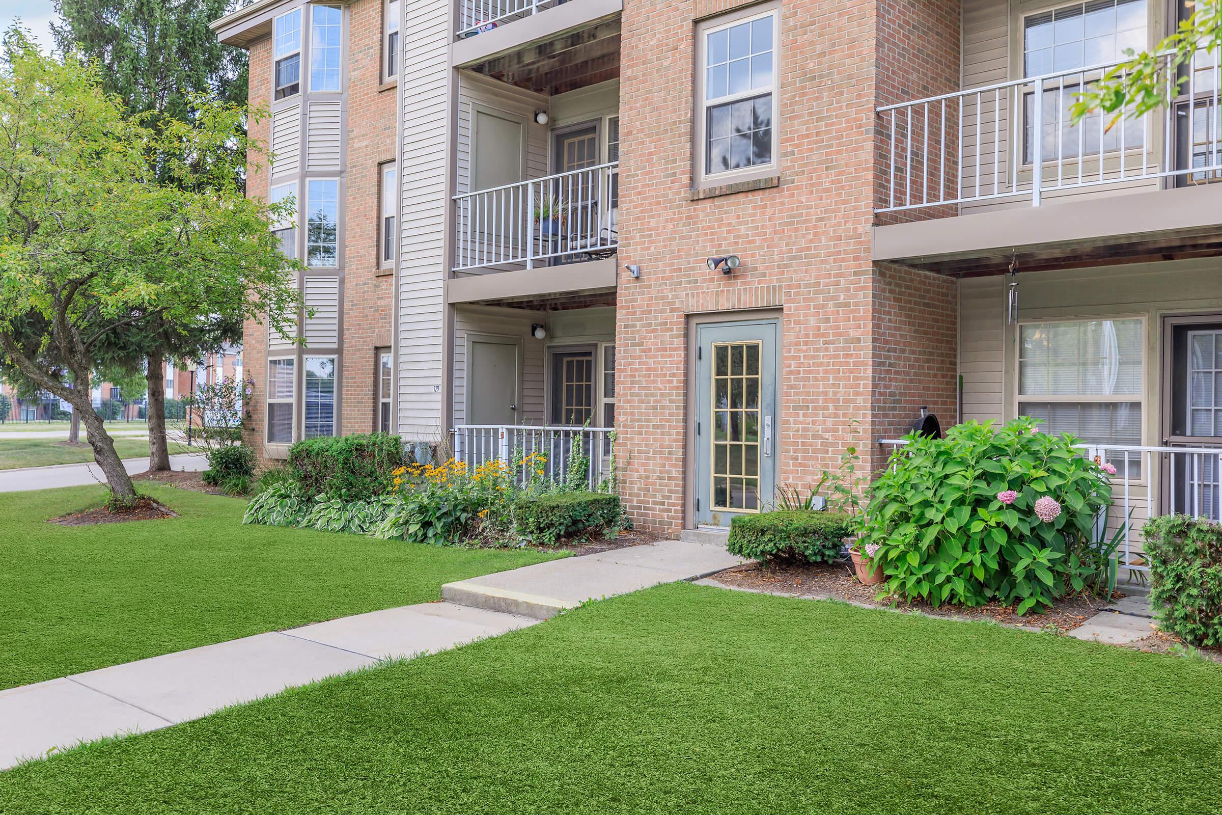 A well-maintained apartment exterior featuring a brick facade, multiple balconies, and a landscaped front yard with flowers and greenery. A concrete walkway leads to the entrance, surrounded by lush grass and decorative plants. The scene is bright and inviting, showcasing a peaceful residential atmosphere.