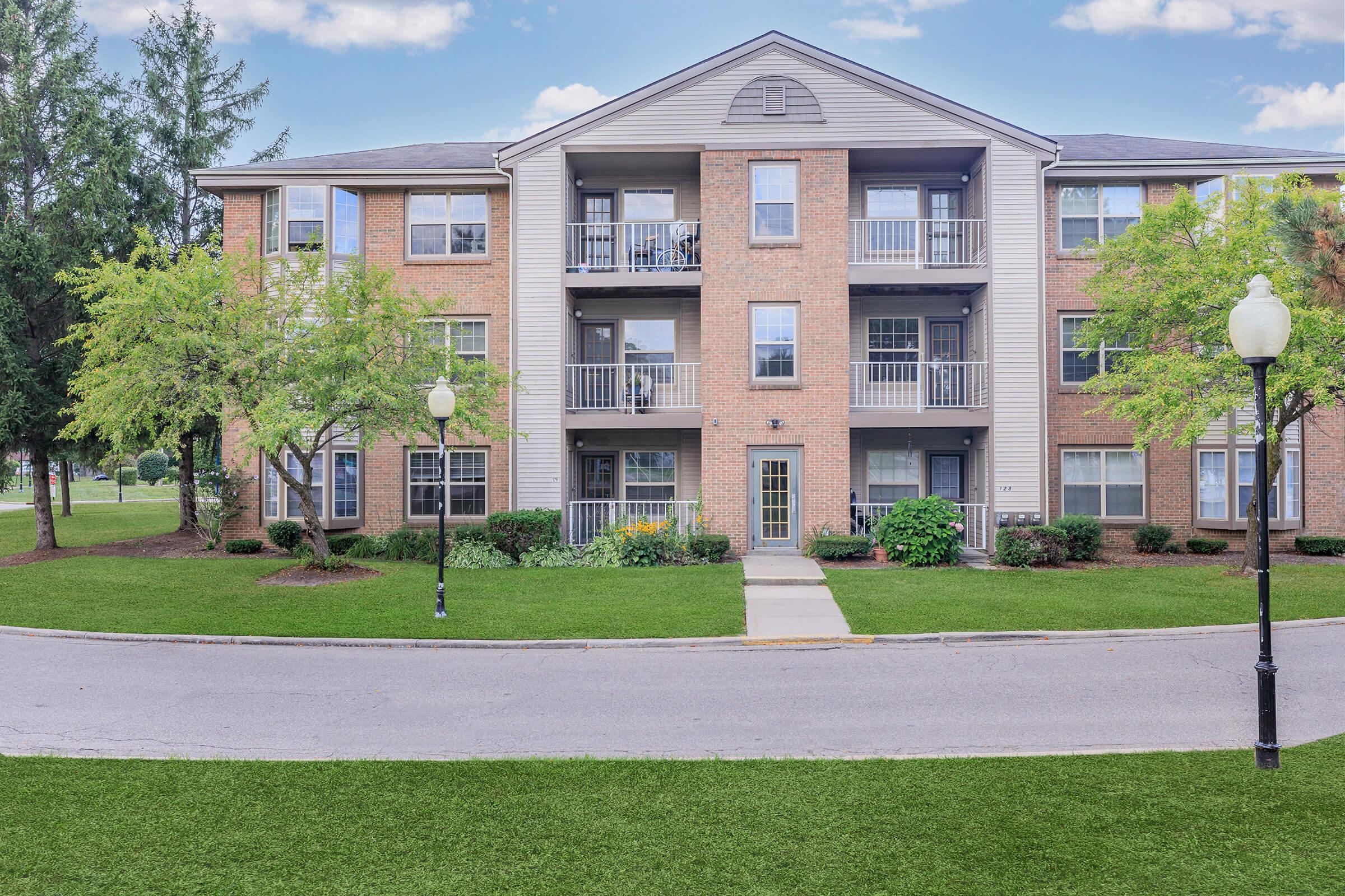 A multi-story apartment building with brick exterior and balconies. Lush green landscaping surrounds the building, including trees and shrubs. A walkway leads to the entrance, and streetlamps line the path. The sky is clear with a few clouds, enhancing the pleasant atmosphere of the scene.