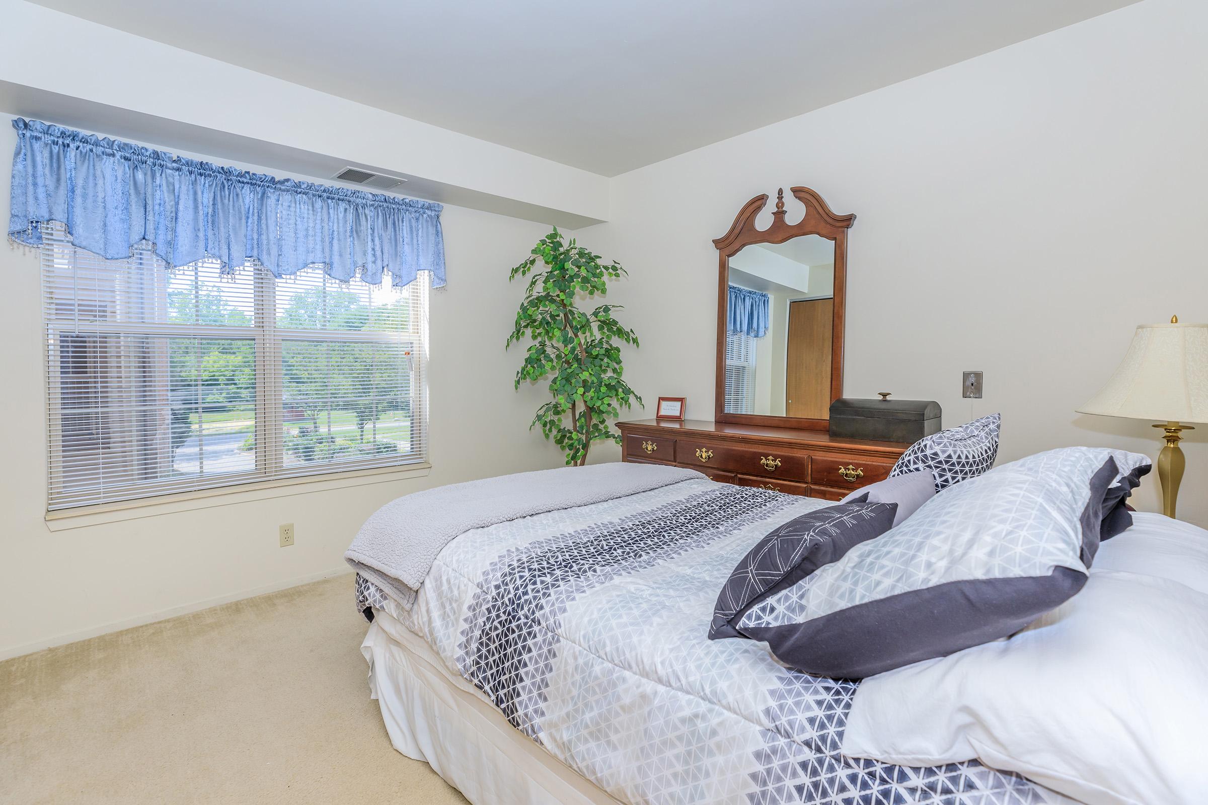 Cozy bedroom featuring a neatly made bed with patterned bedding, a wooden dresser with a mirror, and a large window dressed with blue curtains. A potted plant adds a touch of greenery to the space, and soft lighting creates a welcoming atmosphere.